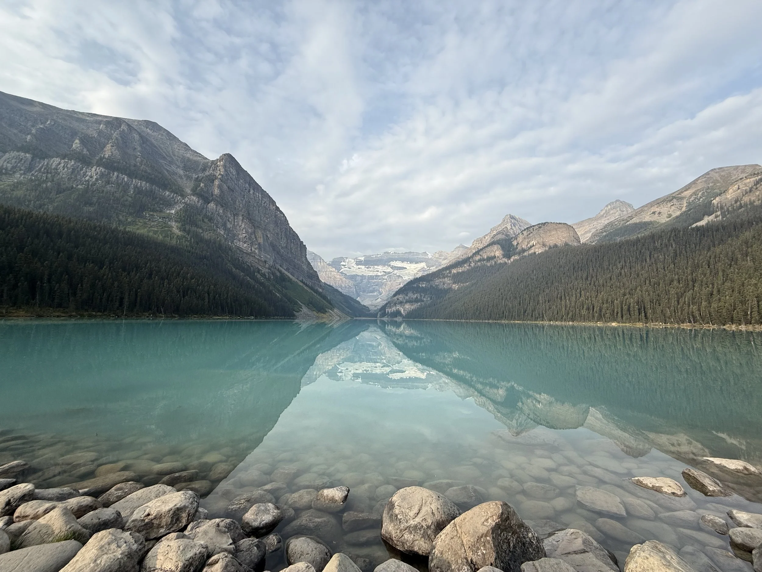 A serene mountain lake with turquoise water, surrounded by tall, forested mountains and a partly cloudy sky.