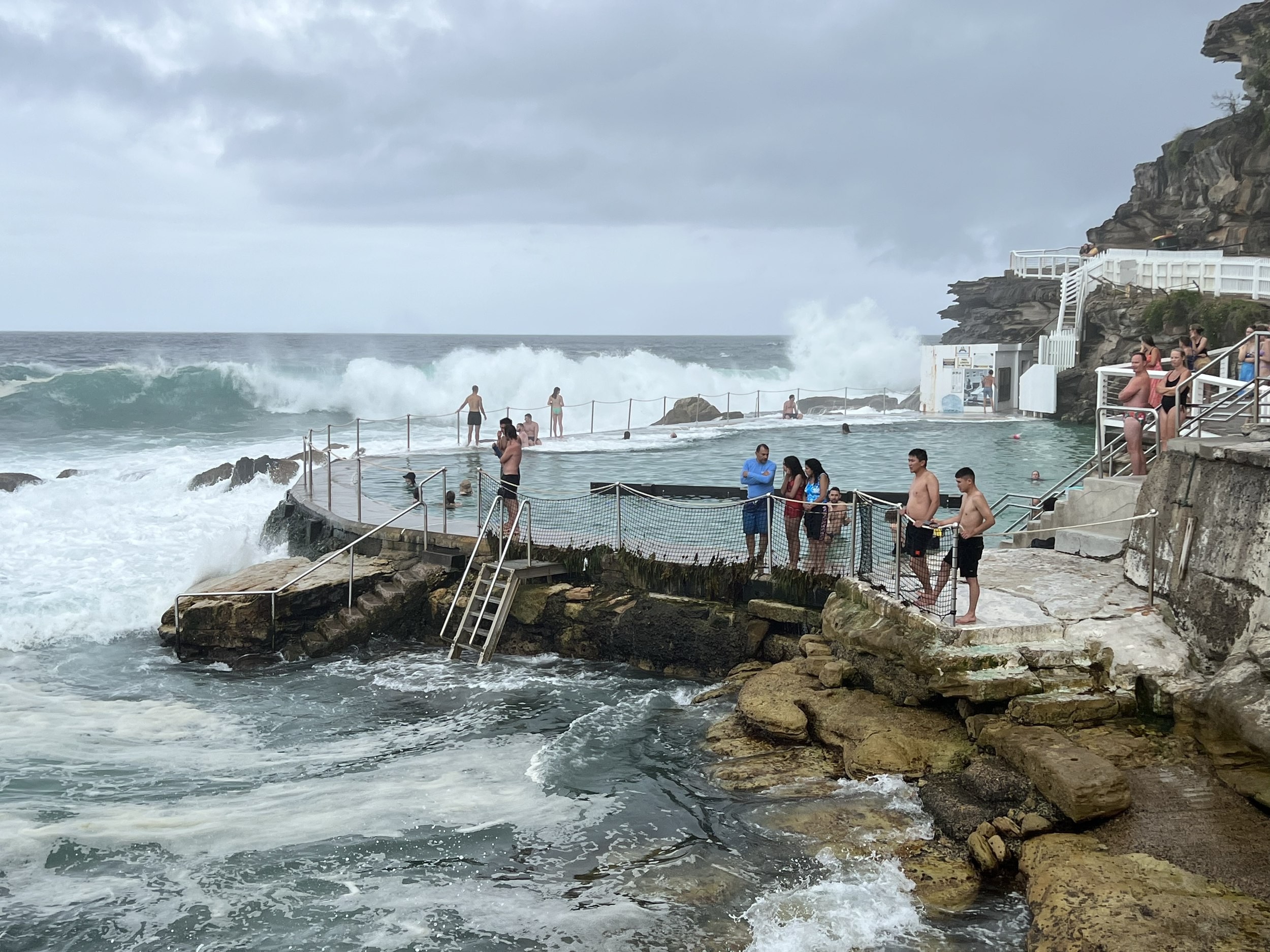 People enjoying the ocean at a seaside hot spring pool, with waves crashing in the background and a cloudy sky overhead.