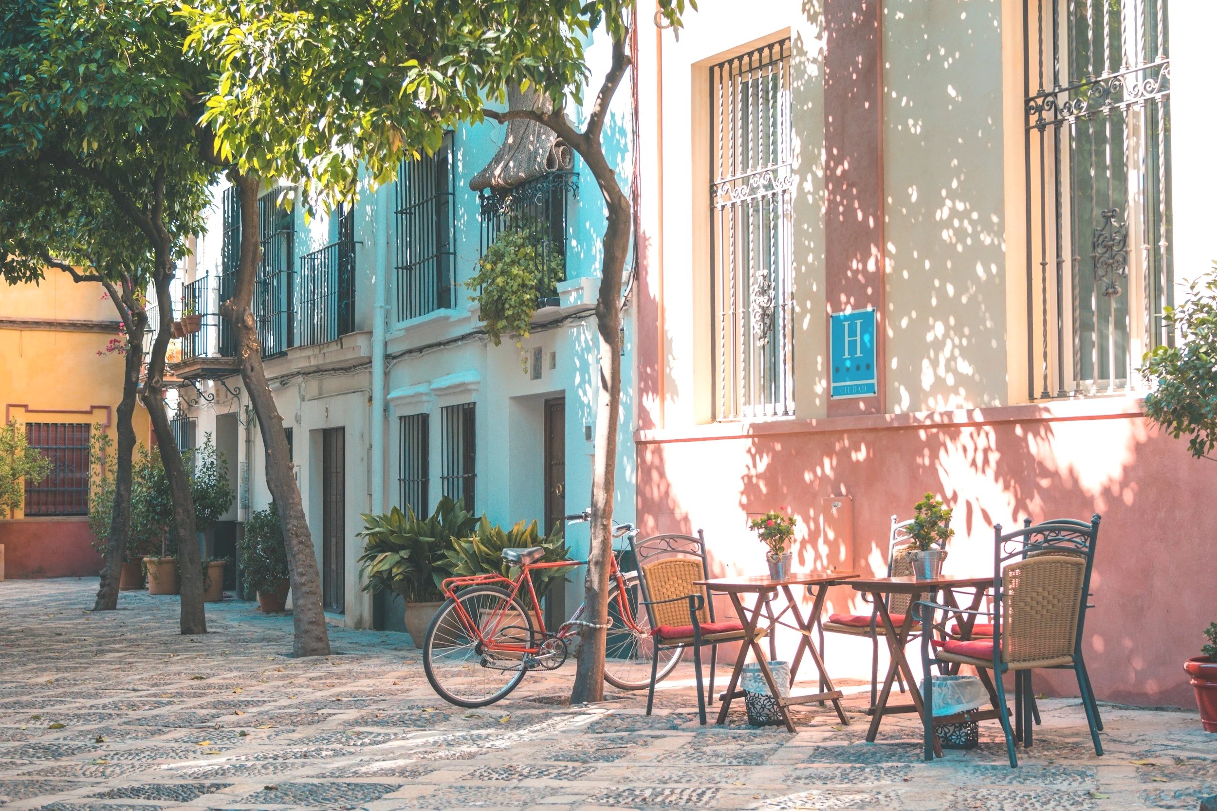 Cobbled street with outdoor café tables, chairs, a pink building, trees, and a bicycle parked next to a tree in a sunny outdoor setting.