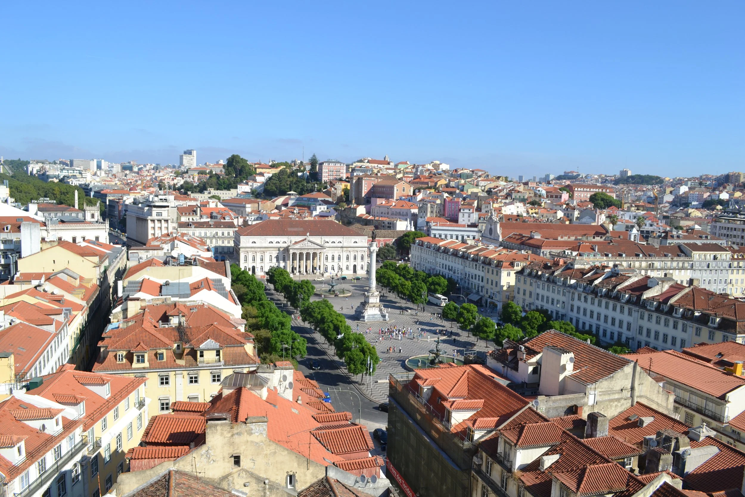 A panoramic view of a city with red-tiled roofs, a large white building, a column monument in the center, and streets with trees and a fountain, under a bright blue sky.