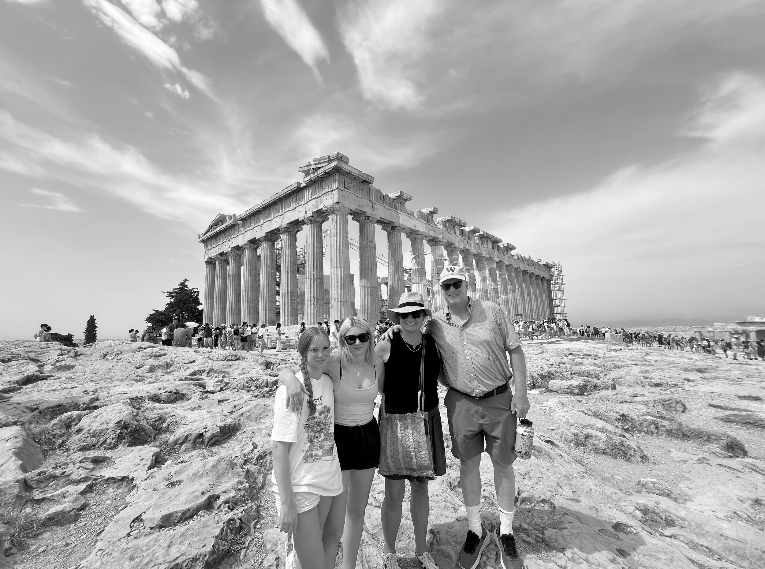 Four people pose in front of the Parthenon on the Acropolis in Greece, with crowds of tourists in the background, under a partly cloudy sky.