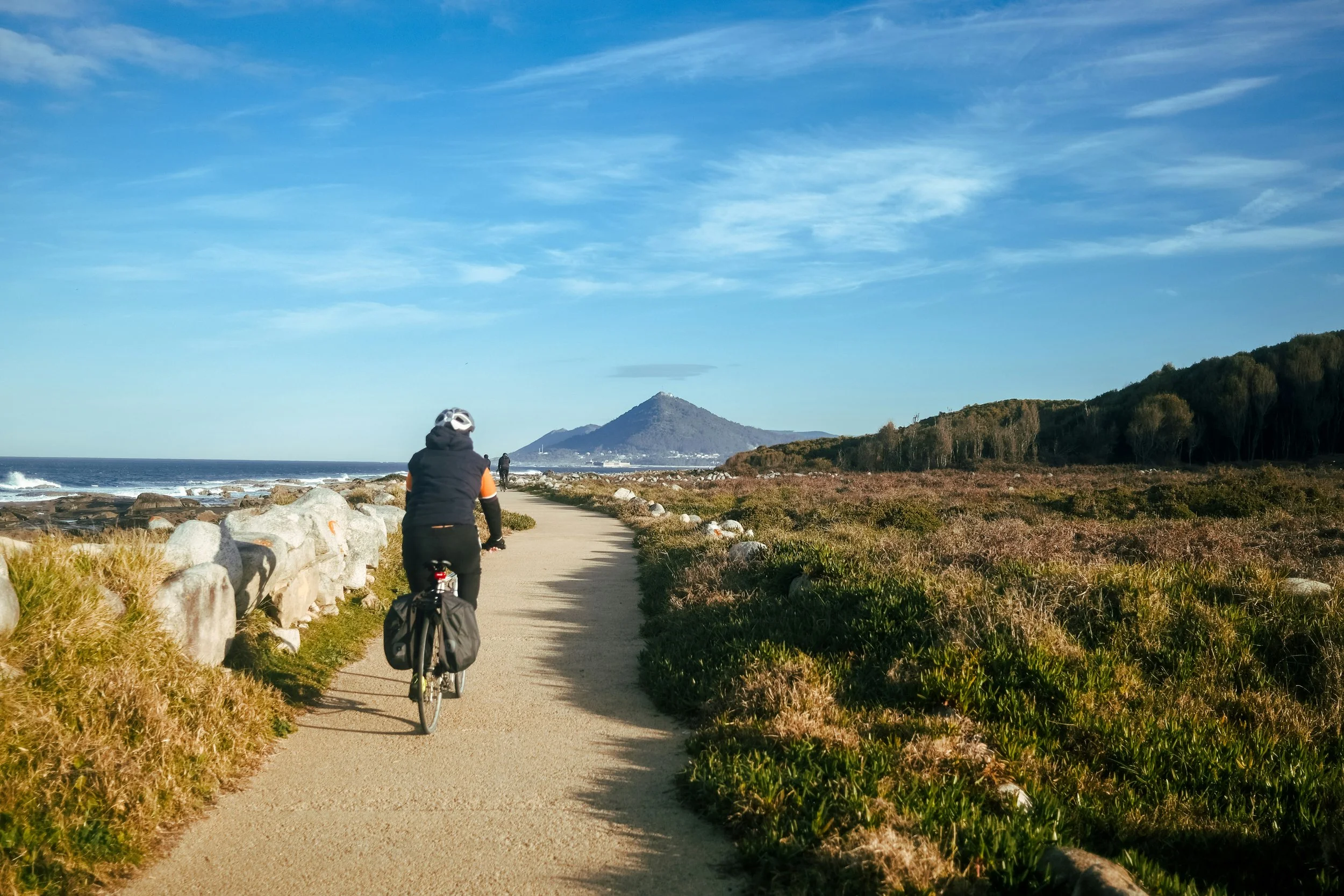 Person riding a bicycle on a coastal trail near the ocean with mountains in the background and a clear blue sky.