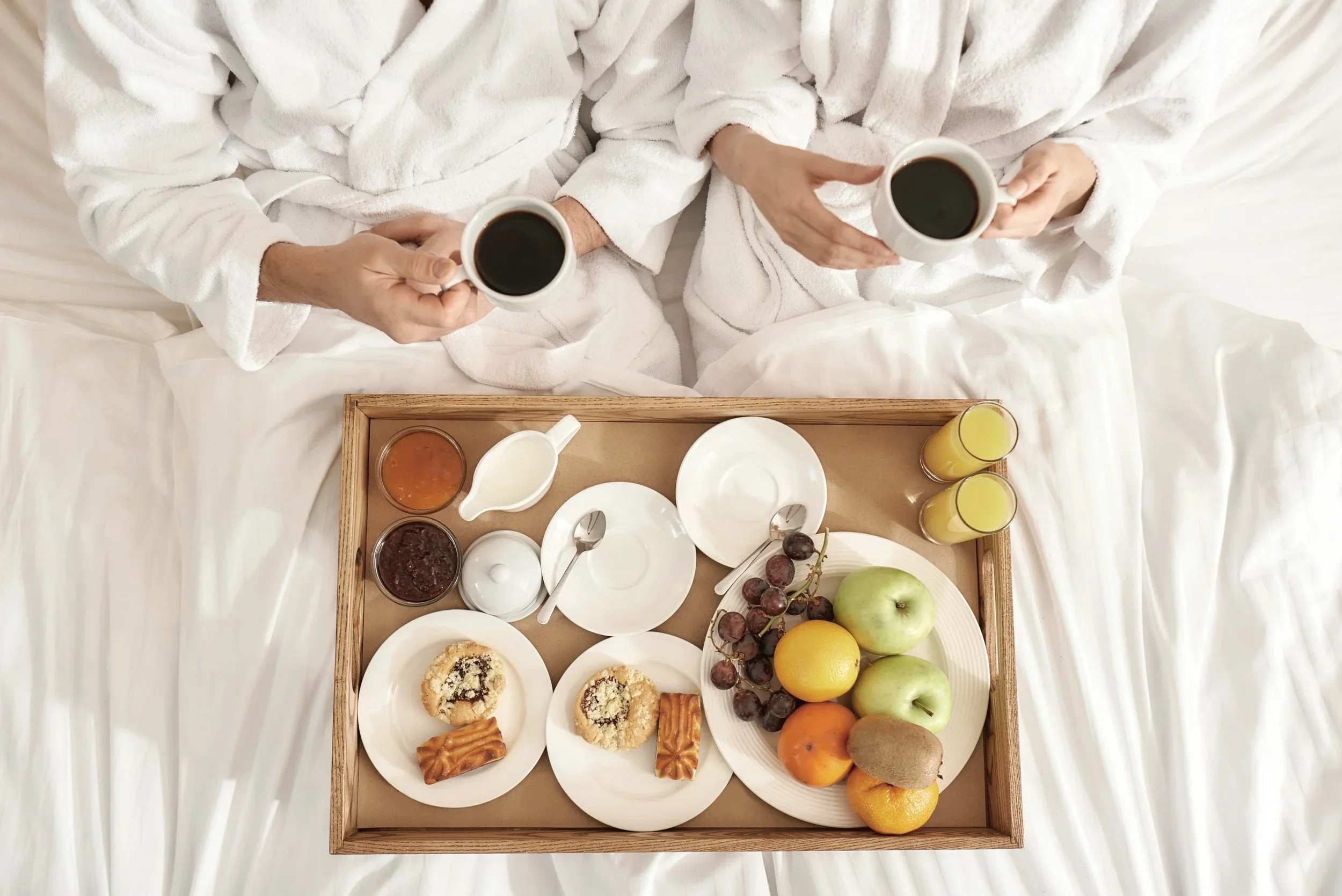 Two people in white robes holding cups of coffee sitting in bed with a tray of breakfast including fruits, pastries, jam, and drinks.