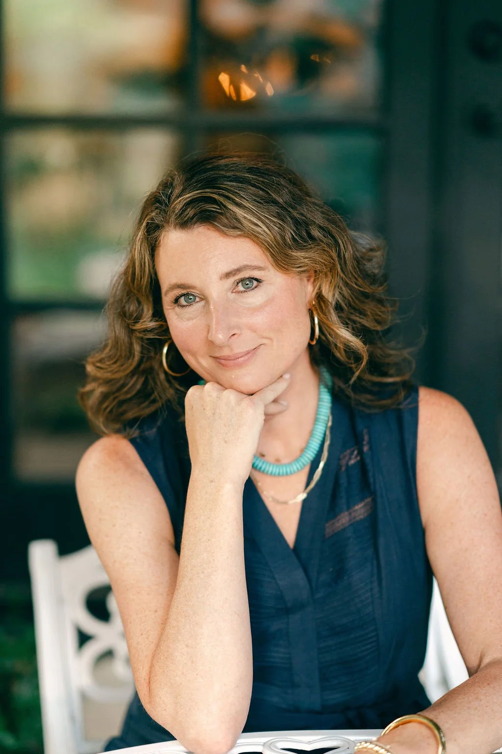 A woman with wavy brown hair, blue eyes, and fair skin, wearing a sleeveless navy top, turquoise necklace, gold hoop earrings, and gold bracelets, resting her chin on her hand and smiling softly at the camera.