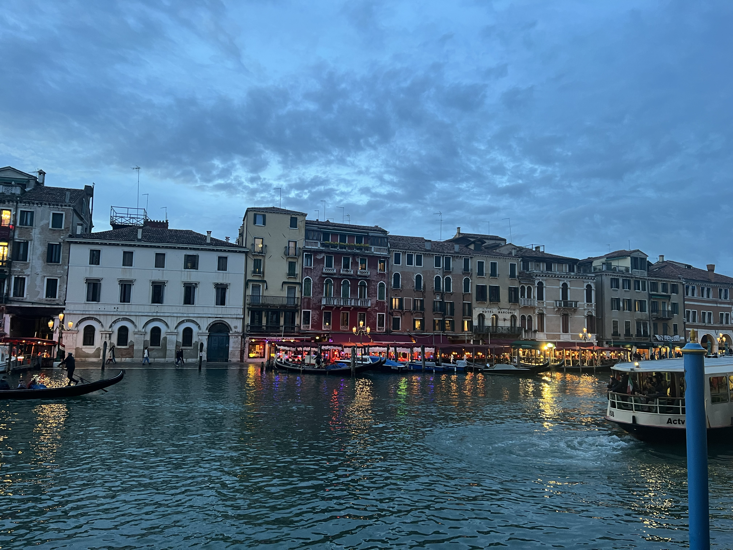 Nighttime view of a Venetian canal with colorful buildings, gondolas, and a water bus, under a cloudy sky.