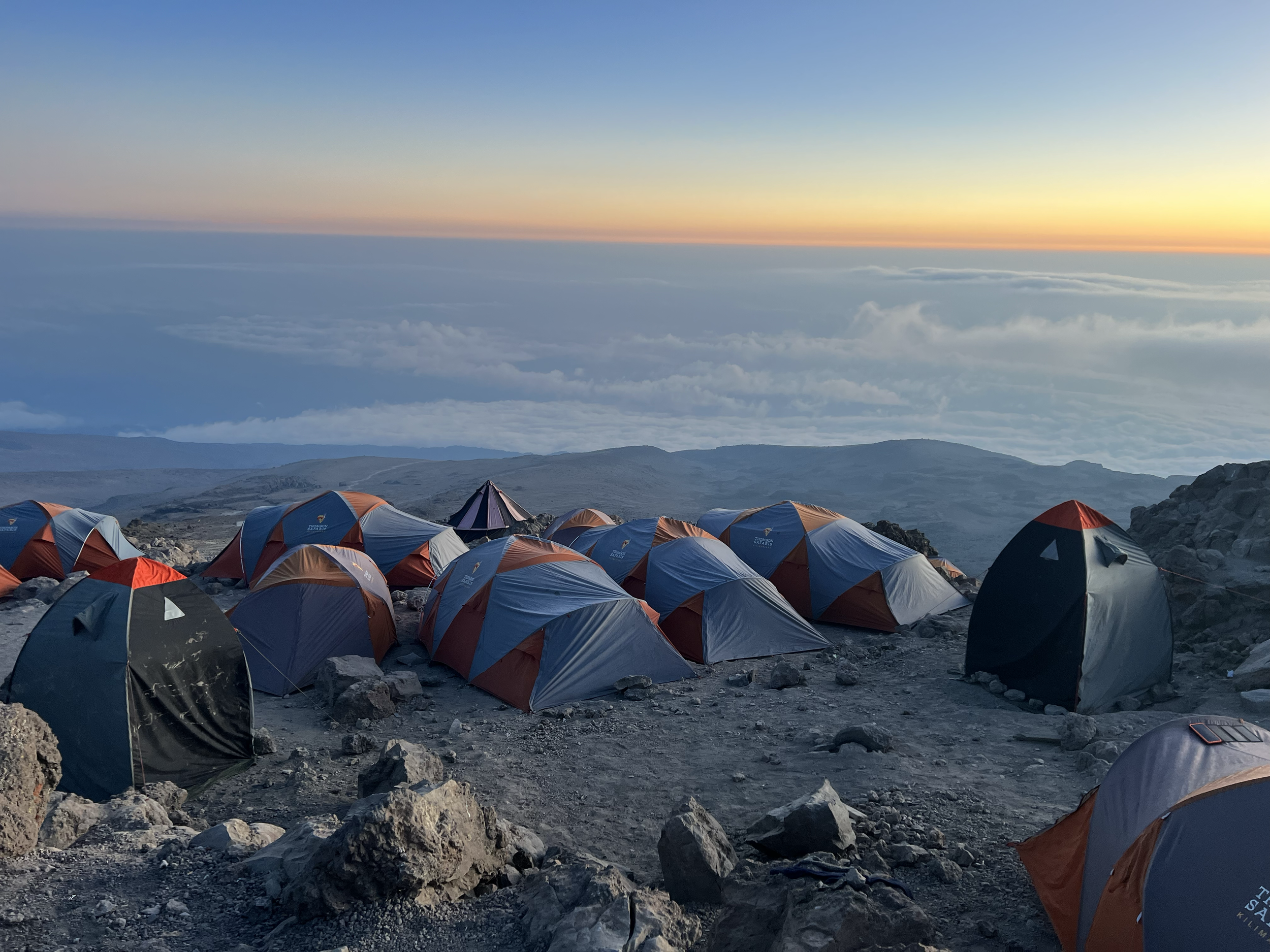 Numerous tents set up on rocky terrain on a mountain during sunset, with clouds and distant mountains in the background.