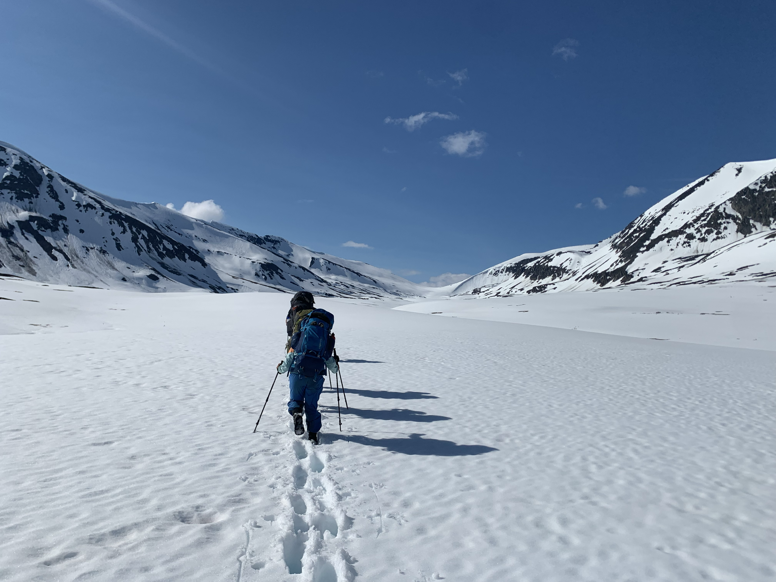 Two hikers with backpacks and trekking poles walking in a snowy mountain valley under a blue sky.