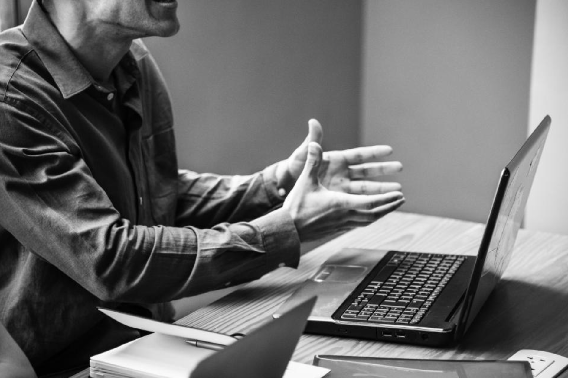 A person wearing a button-up shirt sitting at a desk with an open laptop, gesturing with their hands during a video call or online meeting in black and white.