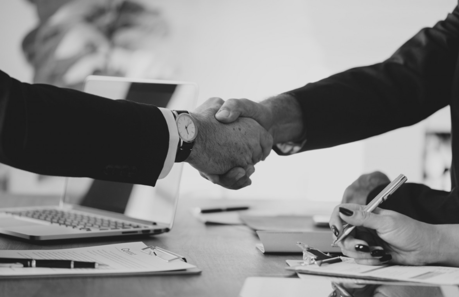 Black and white photo of two people shaking hands across a desk in a business setting. One person is wearing a watch and the other has dark nail polish and is holding a pen.