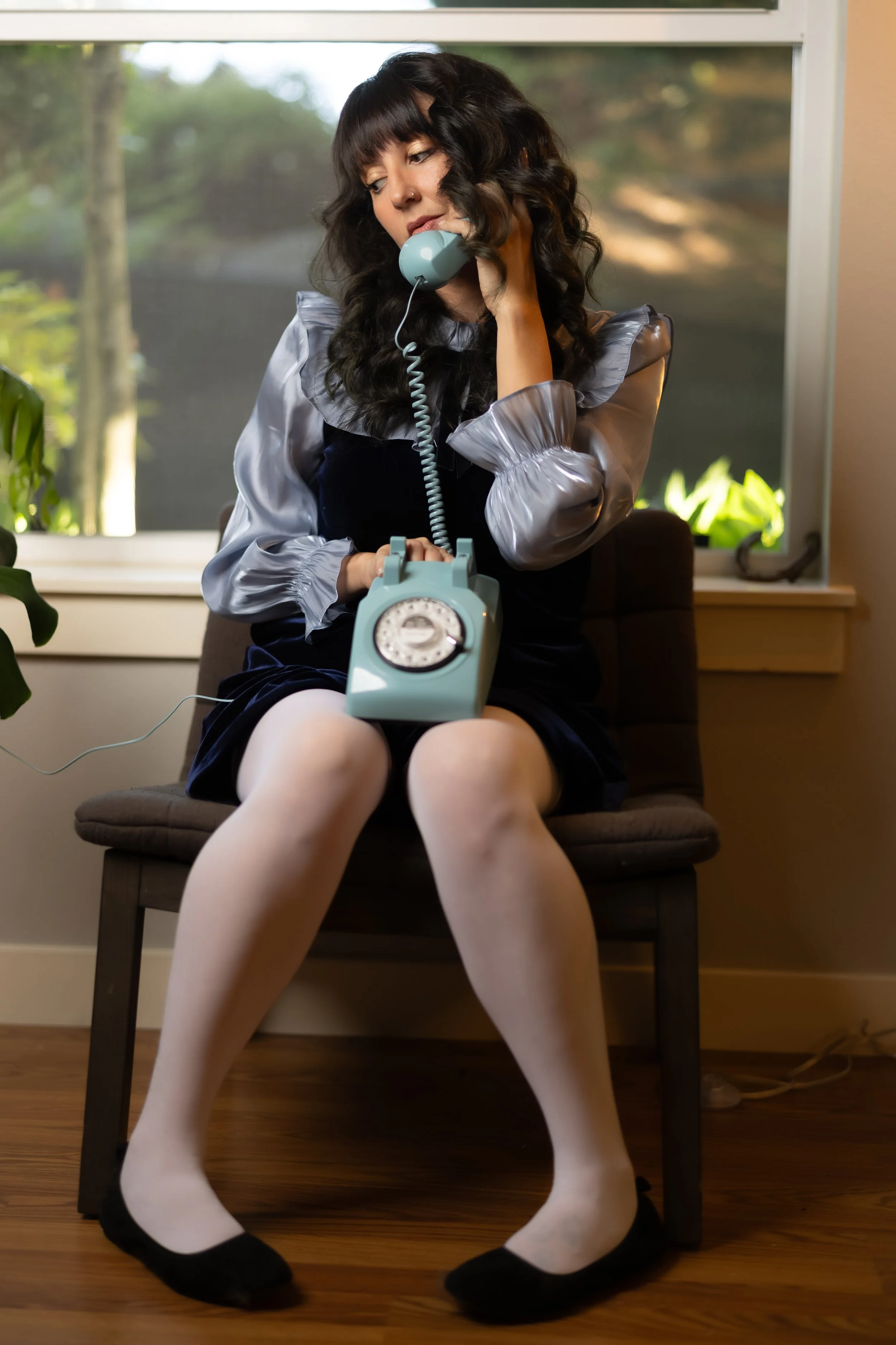 A woman with dark, curly hair and bangs sits on a chair in front of a window, holding an old-fashioned teal rotary phone to her ear. She wears a shiny gray puff-sleeved blouse, a dark velvet dress, white tights, and black ballet flats. Her expression