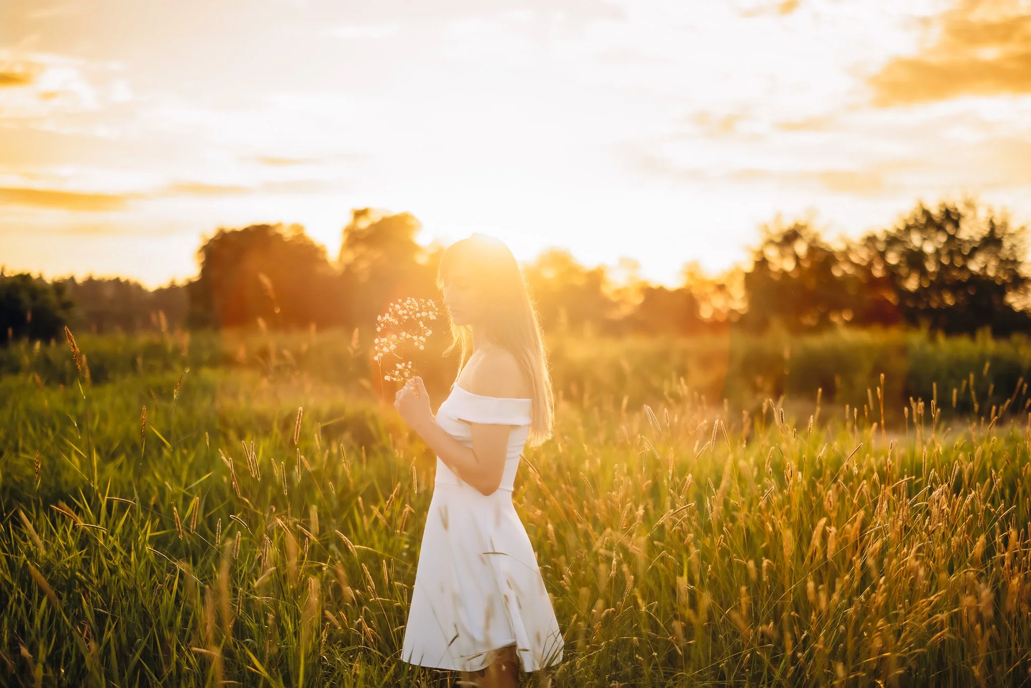 A woman in a white dress standing in a field of tall grass during sunset, holding a small bouquet of flowers.
