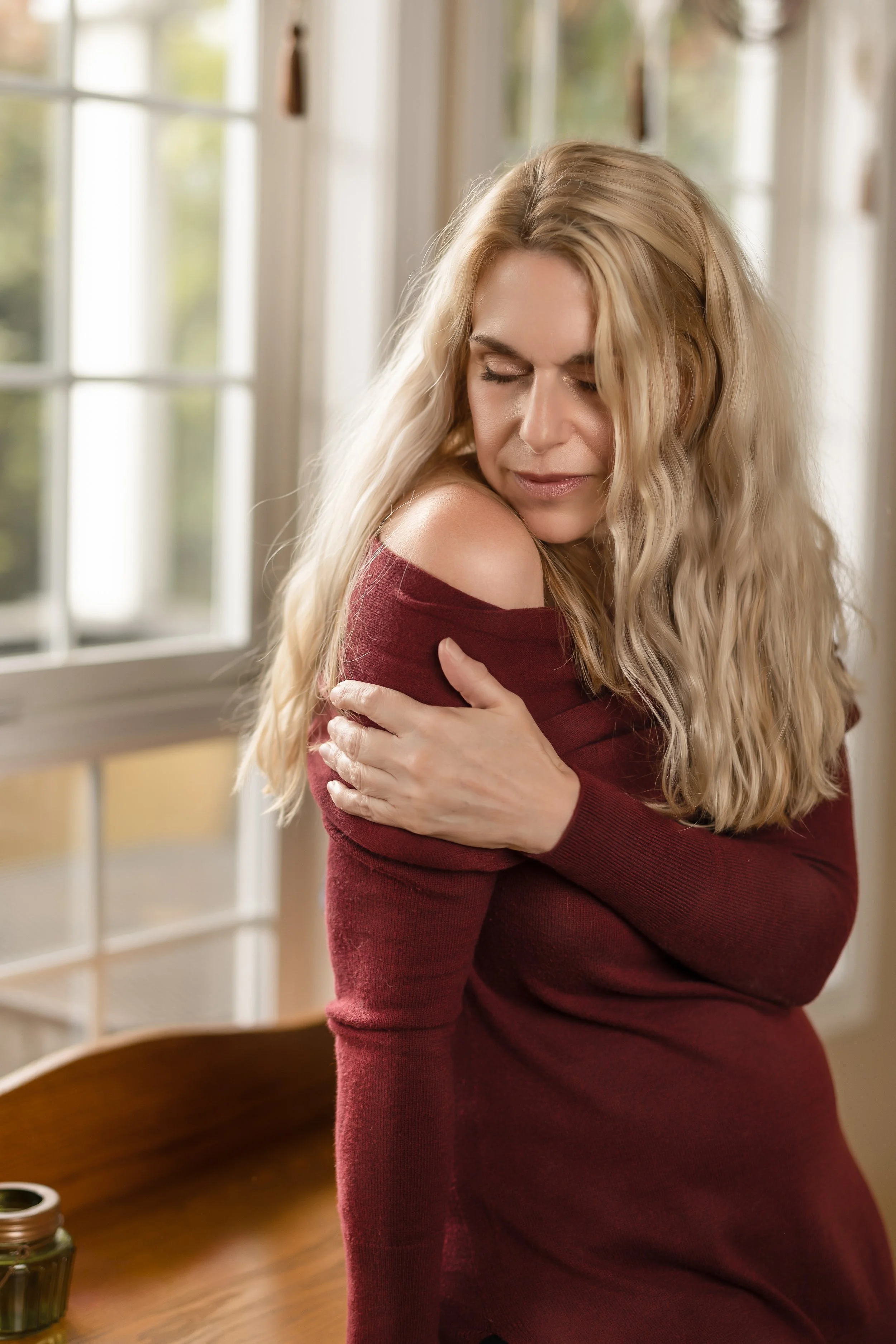 A woman with long blonde wavy hair wearing a reddish-burgundy off-the-shoulder sweater hugging herself, eyes closed, in a room with large windows and a wooden table.