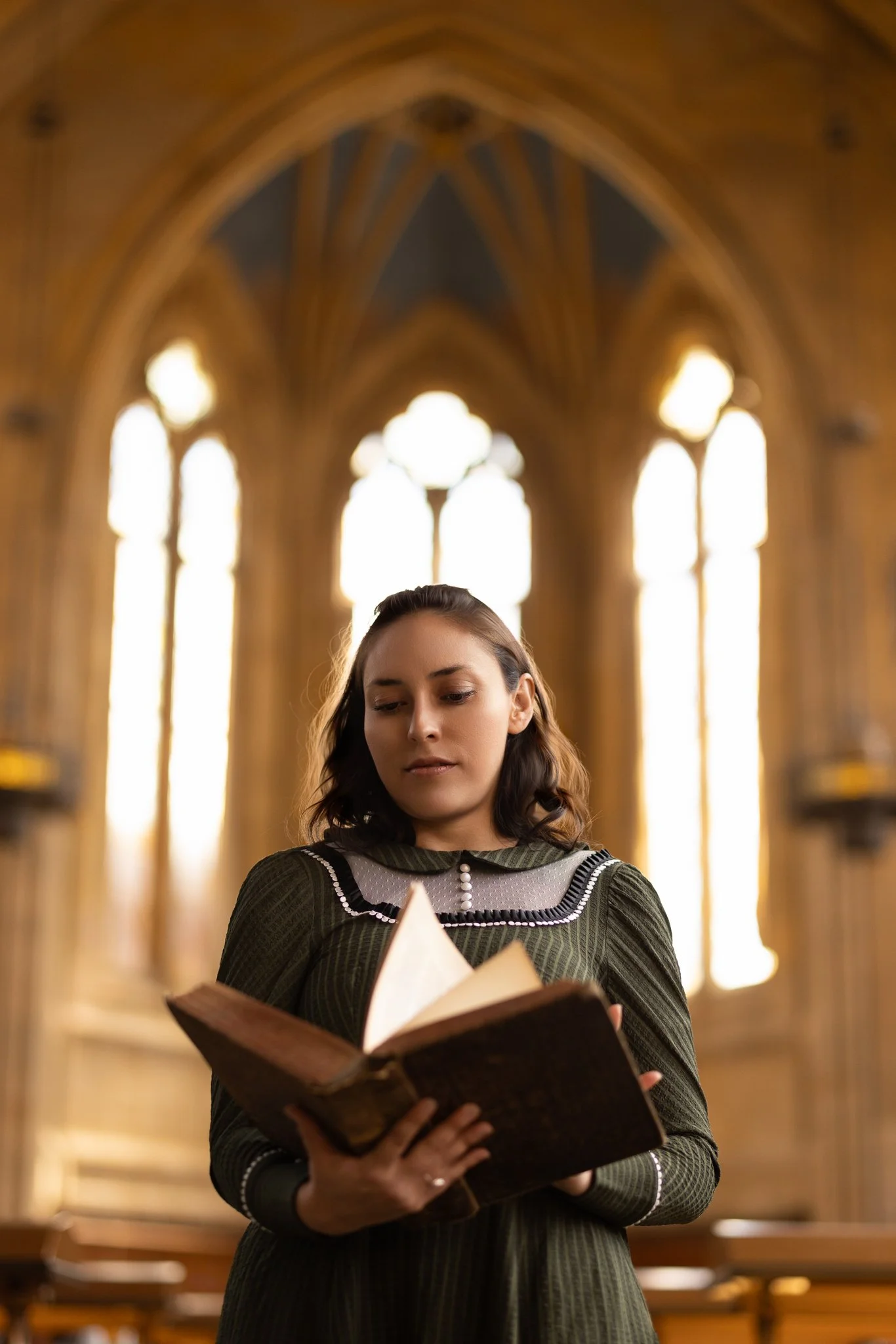 A young woman with brown hair wearing a vintage green dress reads an old book in a grand, gothic-style room with tall, stained glass windows.