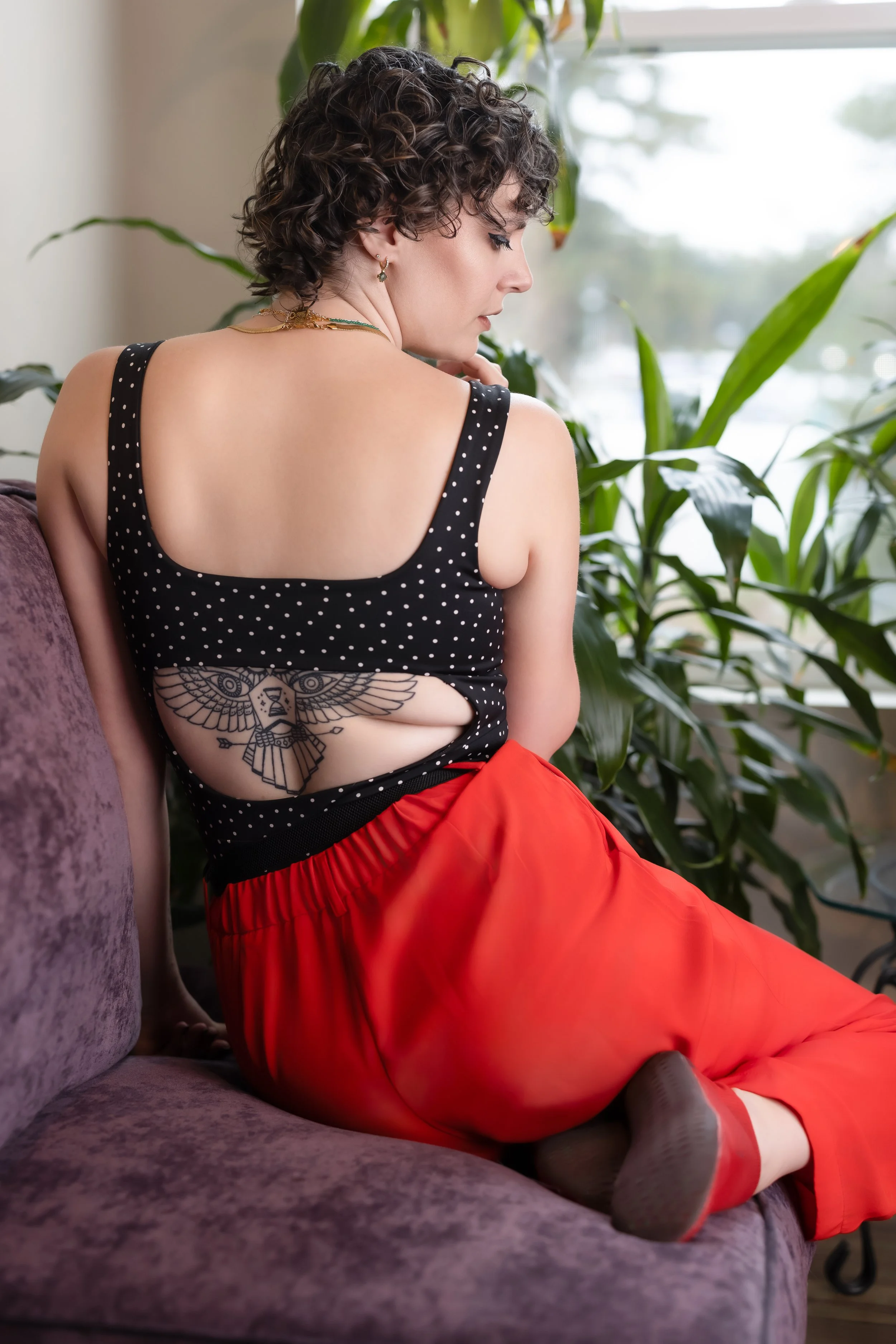 A woman with curly short hair sitting on a purple couch, wearing a black polka dot top with a cutout revealing a tattoo of an owl with outstretched wings on her back, paired with bright orange pants, surrounded by green plants.