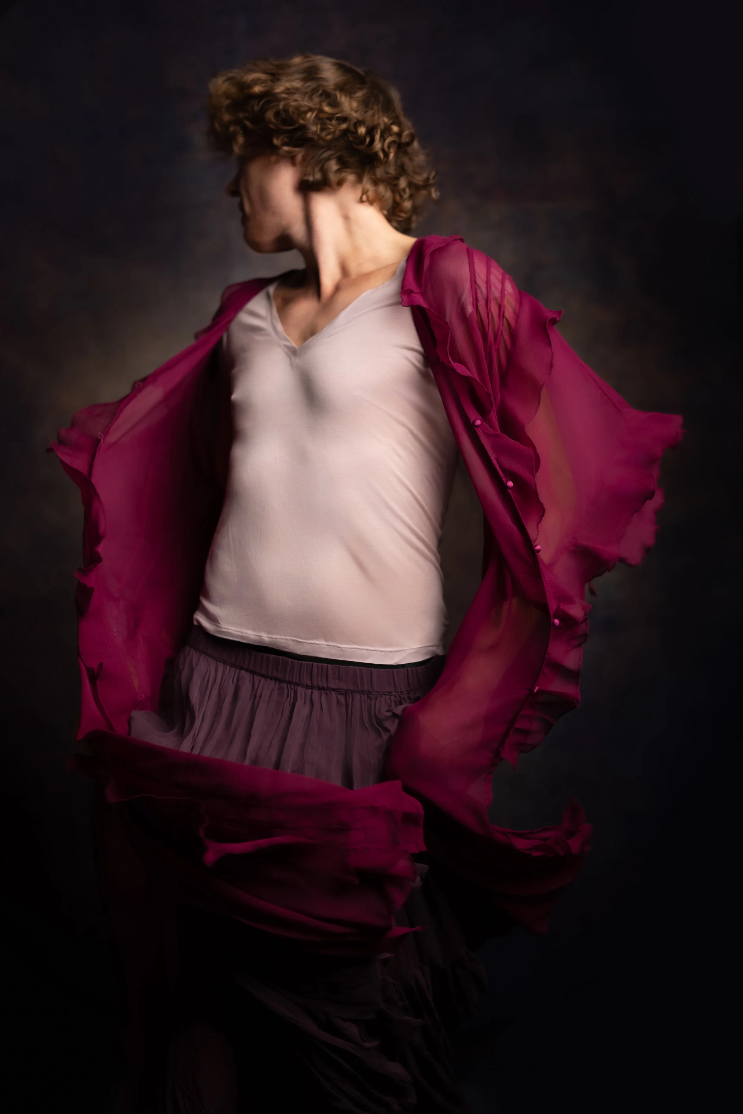 A woman with curly hair in a pink sheer jacket, white top, and dark pants, looking to the side against a dark background.