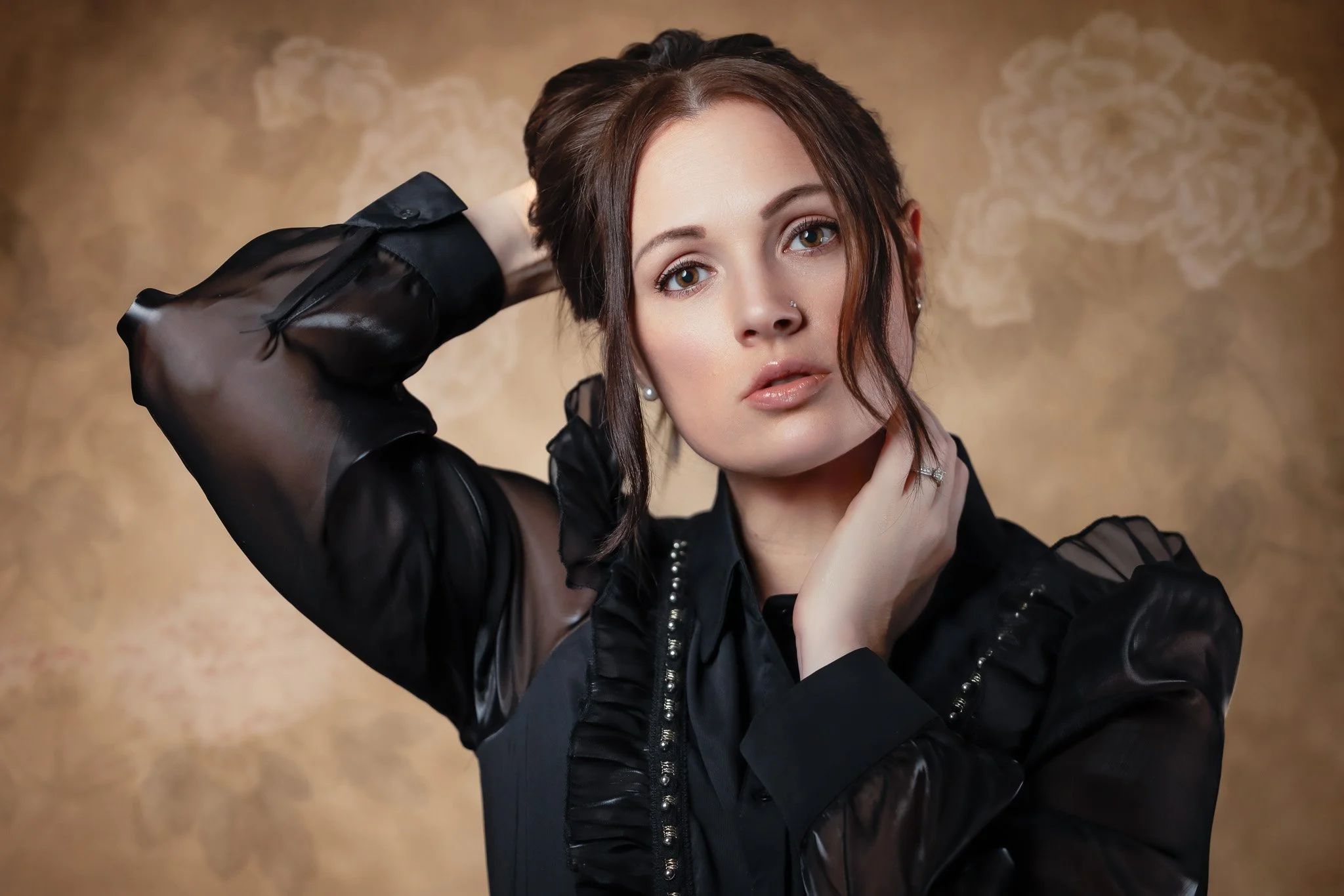 A young woman with brown hair, wearing a black sheer blouse, poses with one hand behind her head and the other touching her neck, against a brown background.
