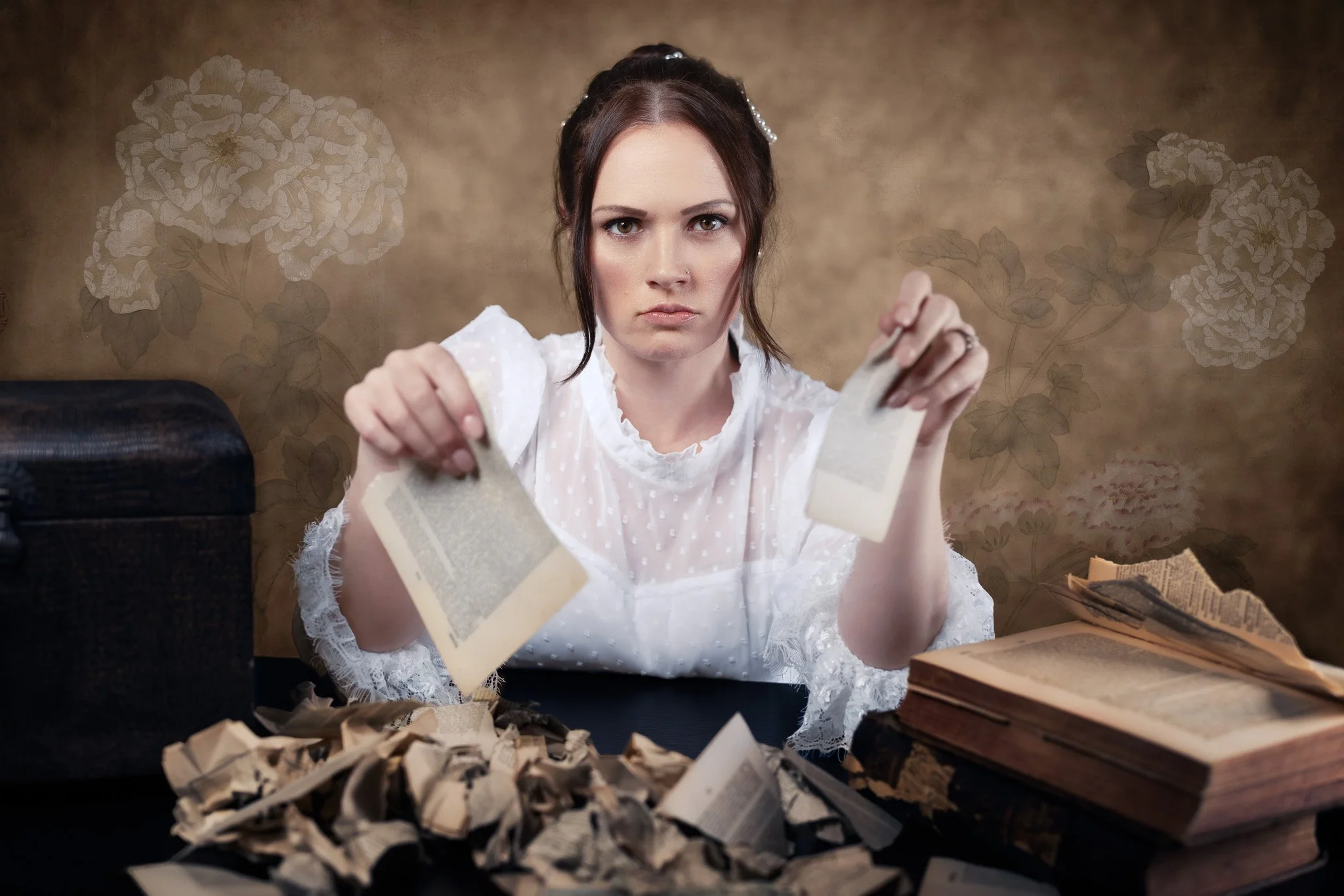 Serious woman with dark hair in a white lace dress tearing paper at a desk surrounded by old books and torn paper, with floral wallpaper in the background.