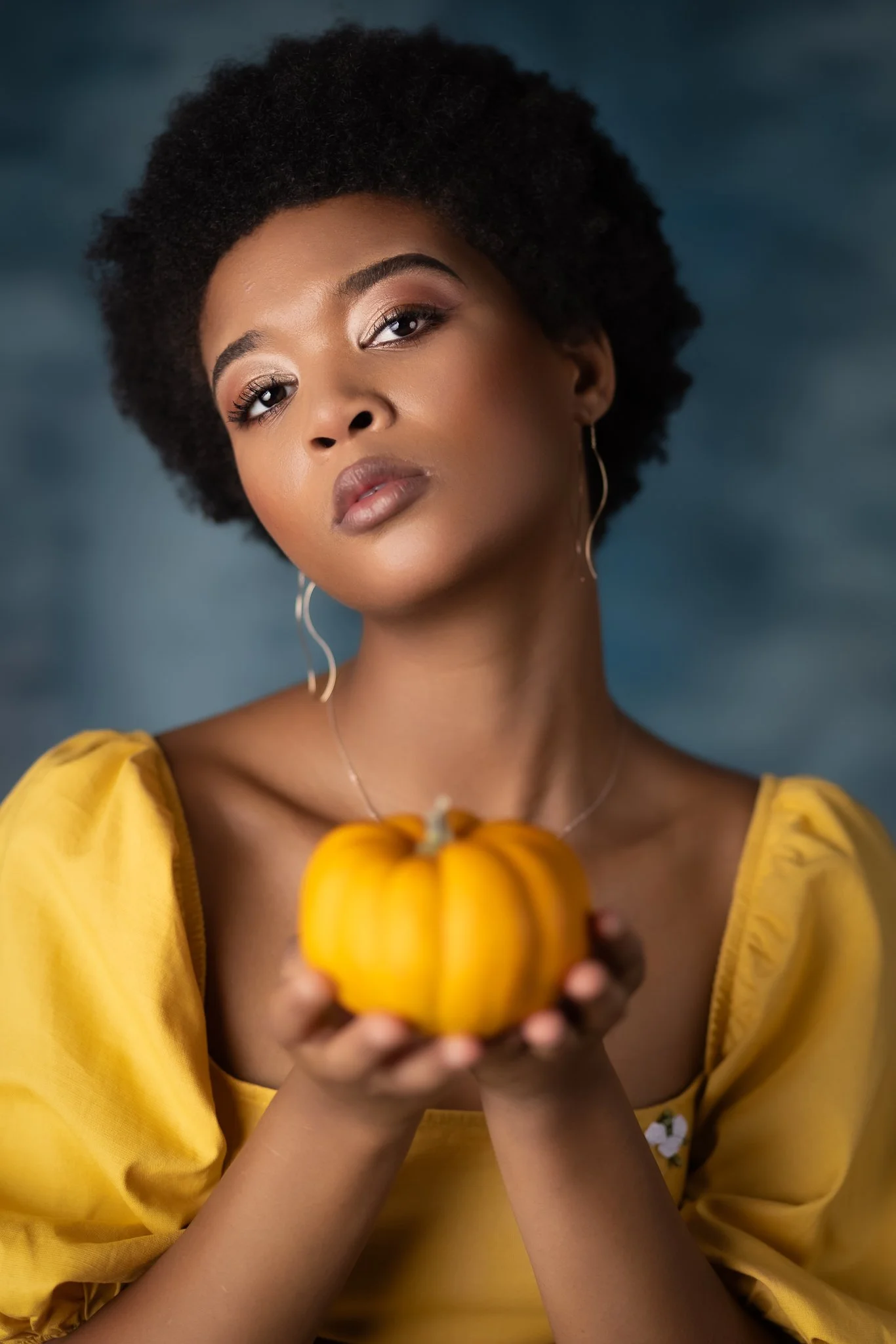 A woman with curly black hair and hoop earrings, wearing a yellow top, holds a small orange pumpkin in her hands, against a dark, cloudy background.
