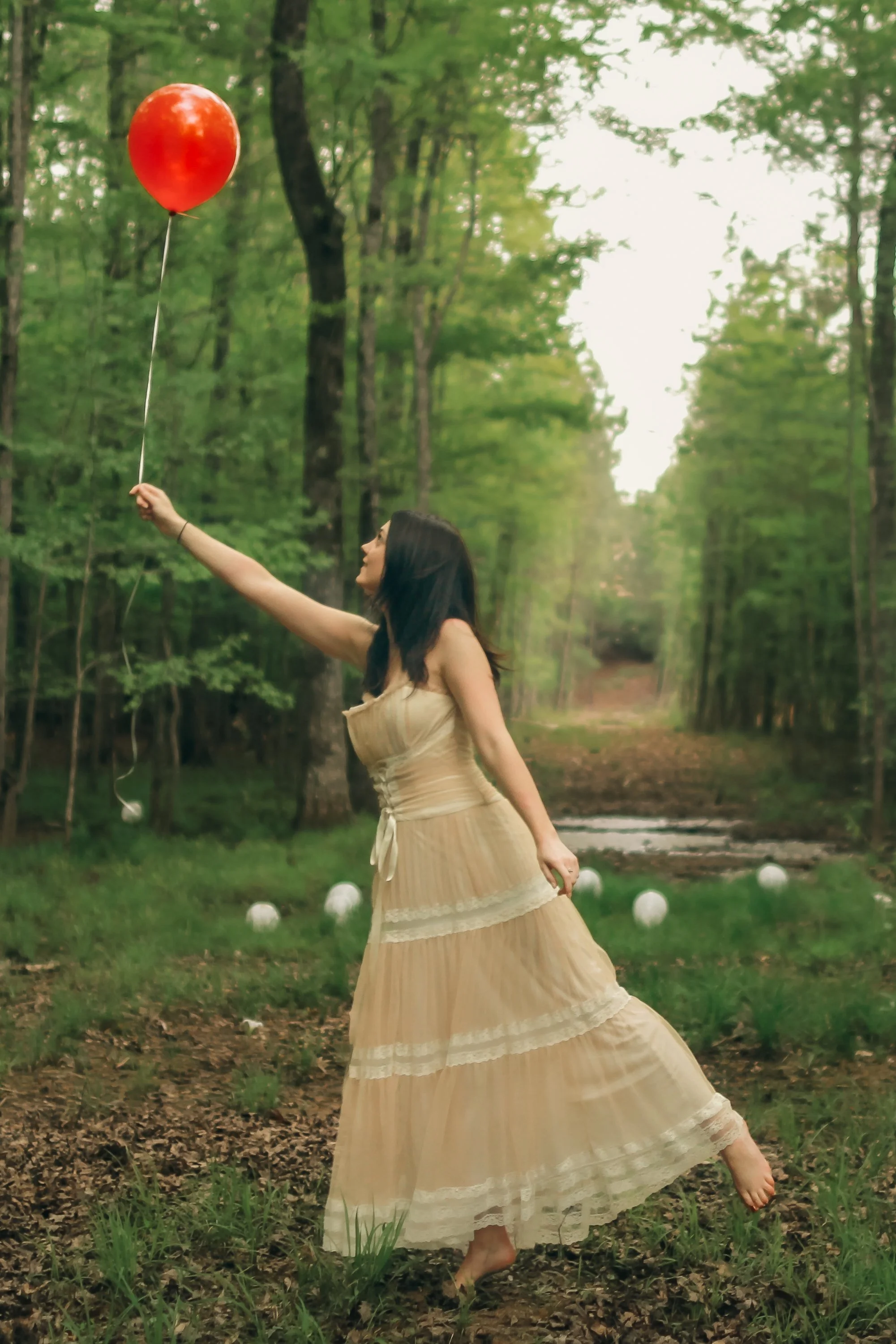 Woman in a beige dress holding a red balloon in a forest clearing with white balloons on the ground