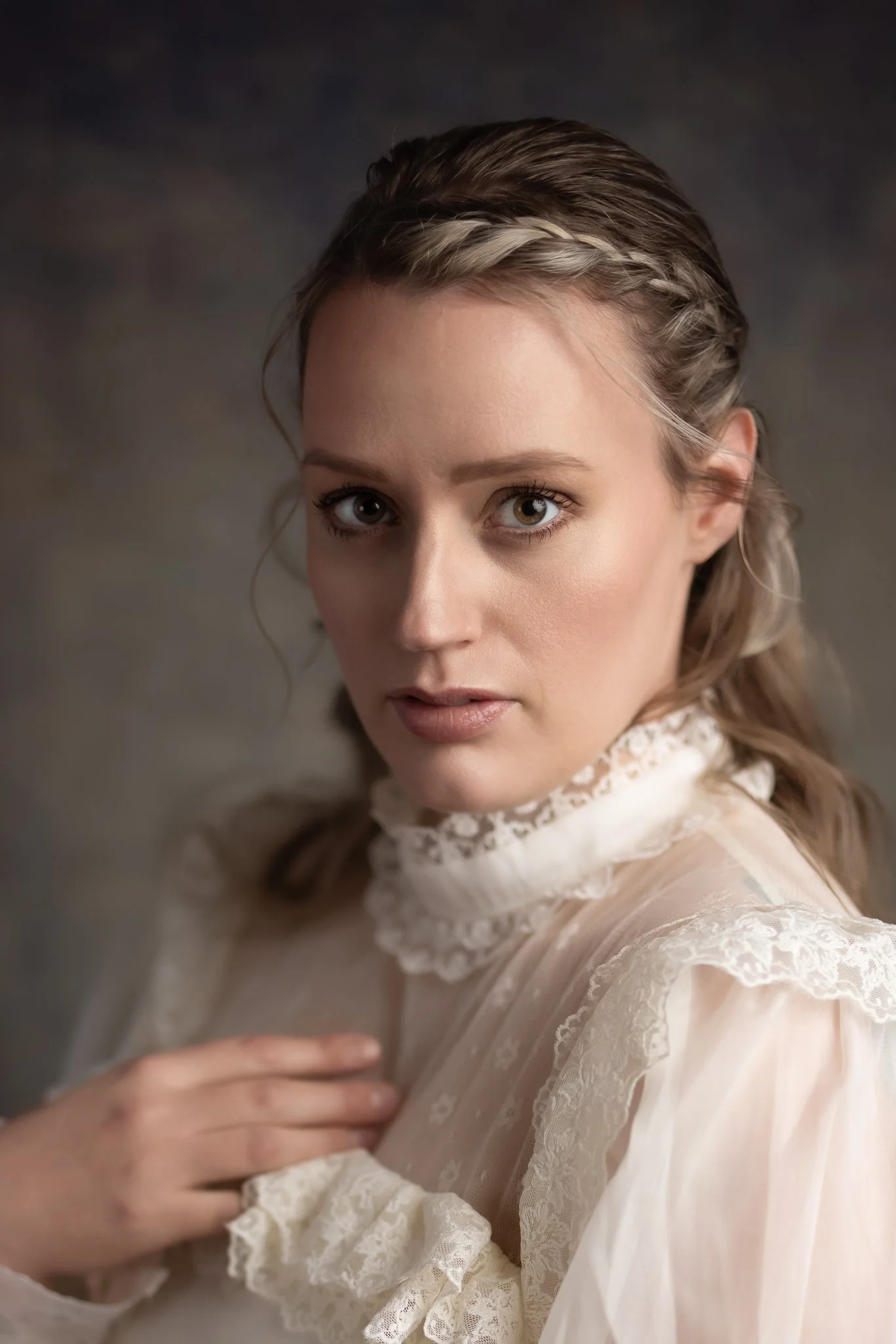 Close-up portrait of a woman with light brown hair styled in soft waves and a braid, wearing a vintage-inspired lace blouse, looking directly at the camera with a neutral expression.
