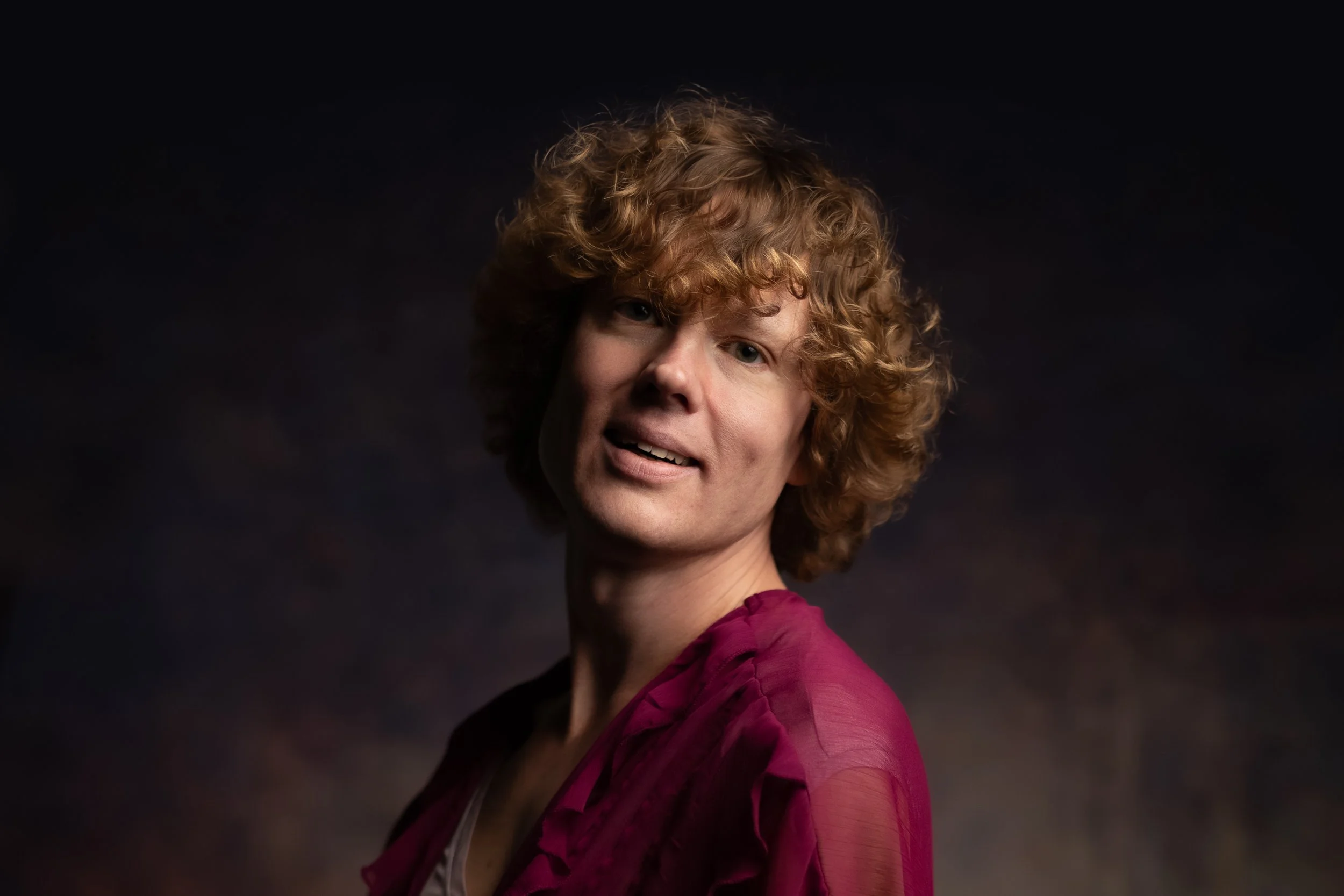 A woman with curly red hair looking at the camera against a dark background.