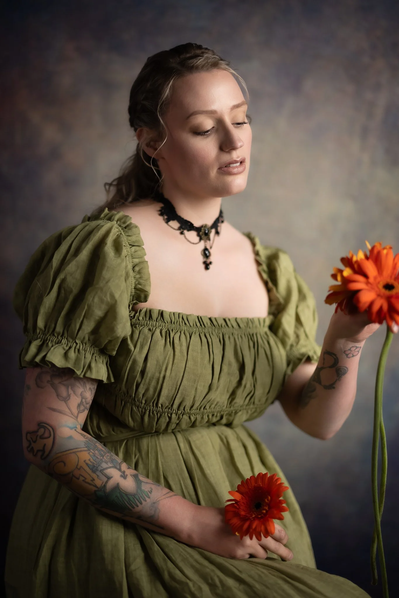 A woman with tattoos on her arms, wearing a vintage green dress with puffed sleeves, holds two orange flowers, one in each hand, while looking down at the flowers.