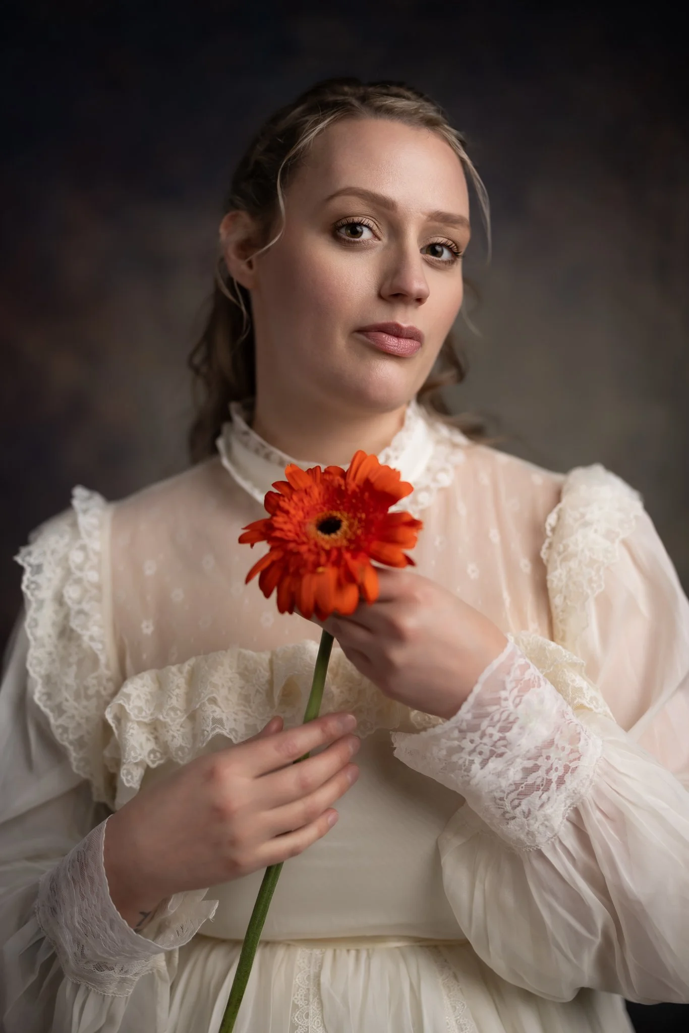 A woman in vintage clothing holding an orange flower against a dark background.