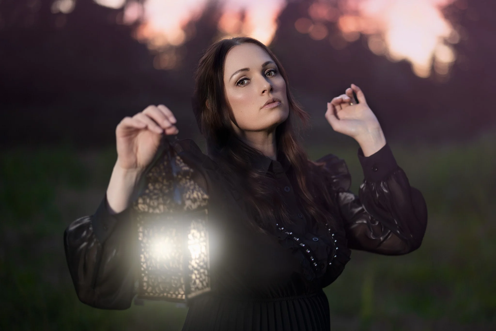 A woman with long dark hair wearing a black lace top and holding a lantern outdoors during sunset.