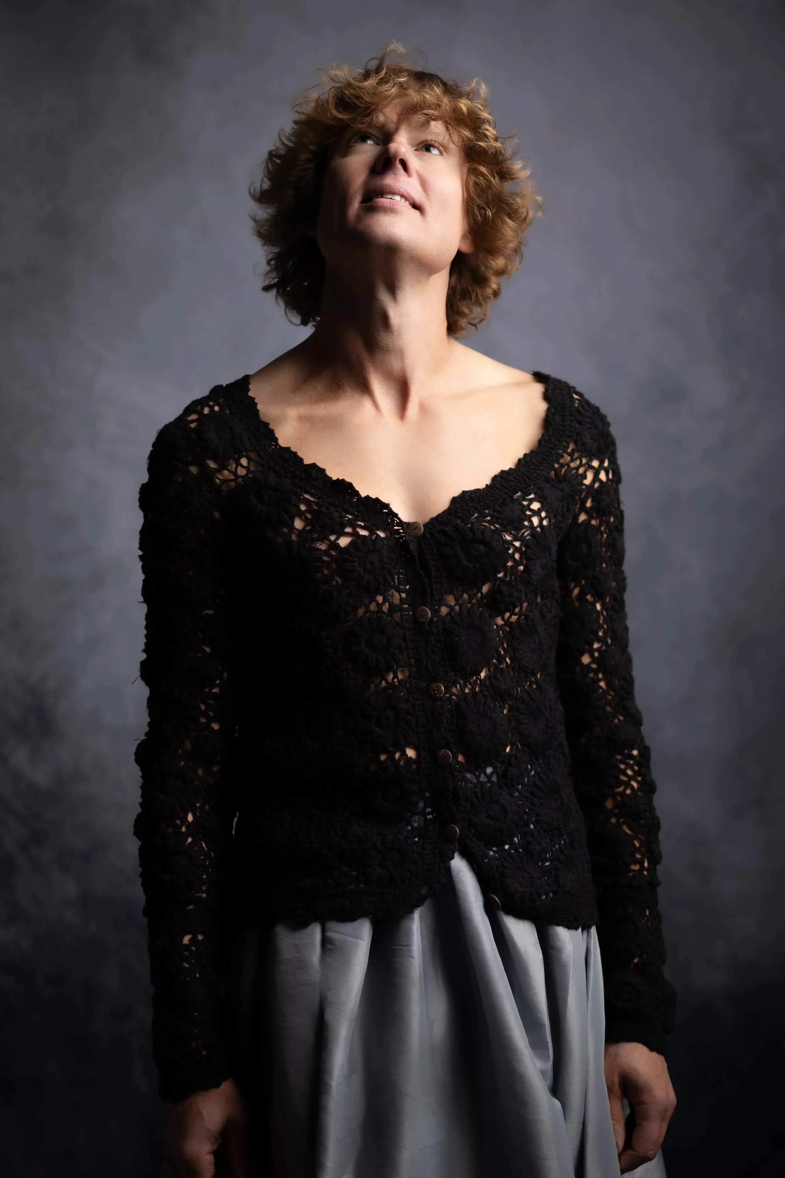 A woman with curly red hair wearing a black lace cardigan and a satin skirt looks upward with a thoughtful expression.