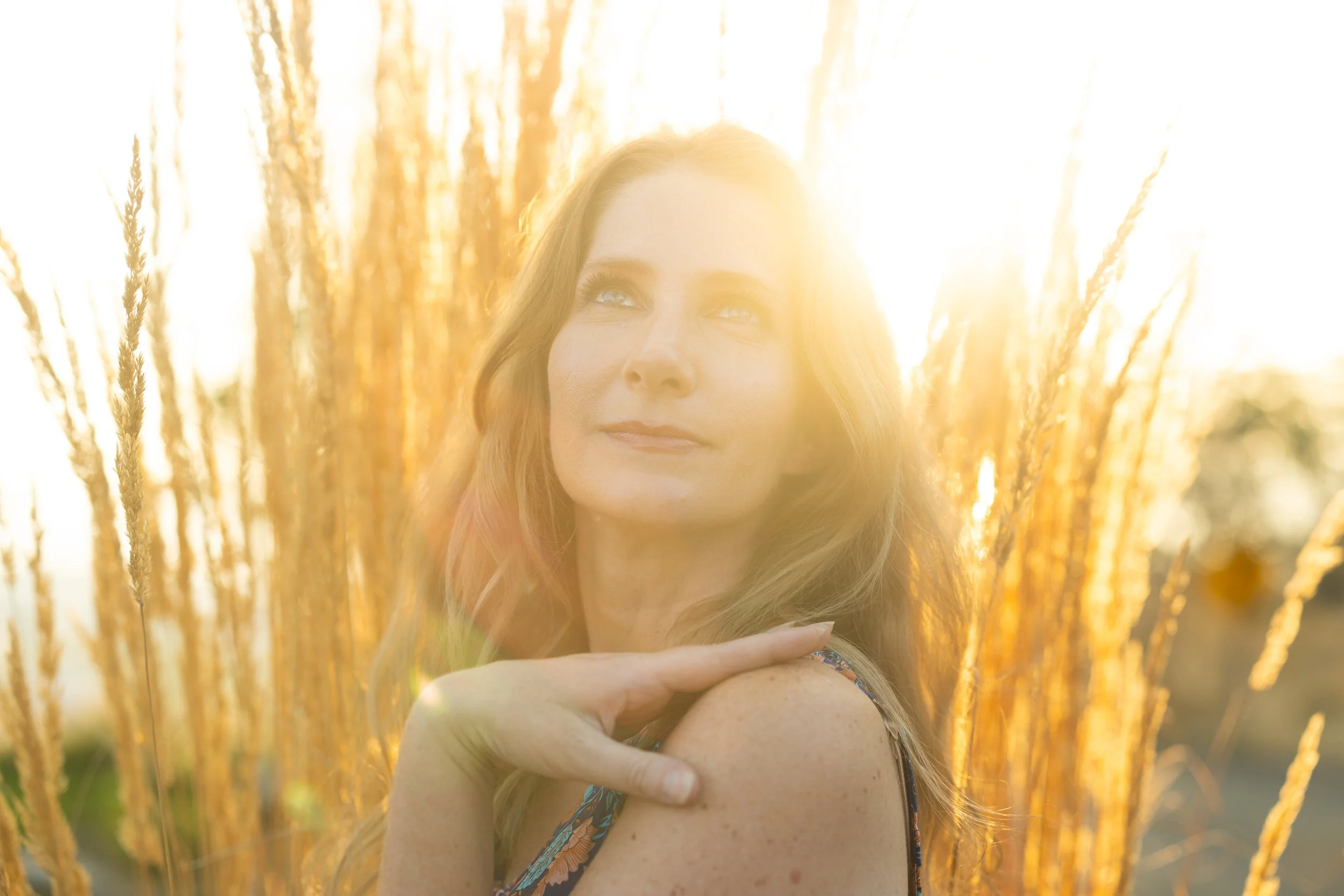 A woman with long, wavy hair standing among tall, golden grasses during sunset, looking slightly upward with a peaceful expression.