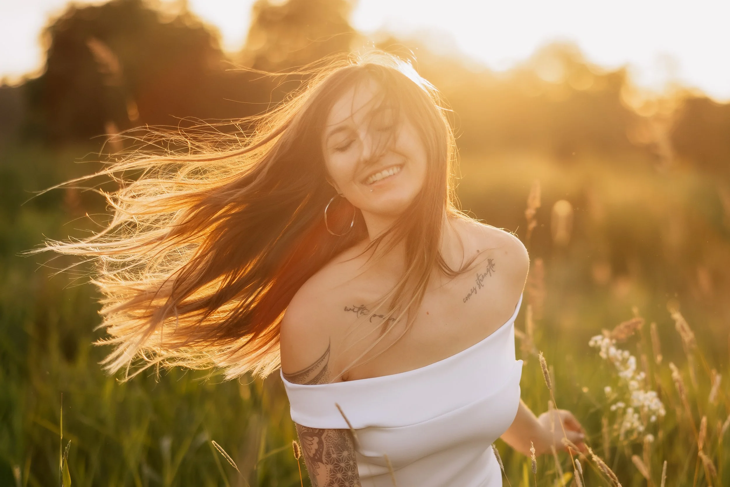 A woman with long hair and tattoos smiling and dancing in a field during sunset.