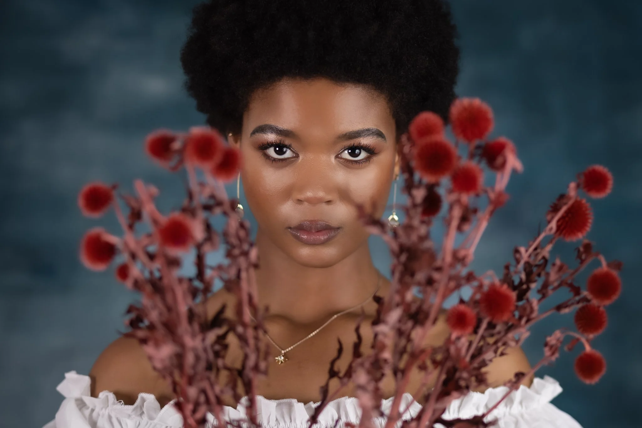 A woman with dark skin, styled in a natural afro, looking directly at the camera, holding a bouquet of red, fluffy flowers. She is wearing a white off-shoulder top with ruffled details, earrings, and a delicate necklace.