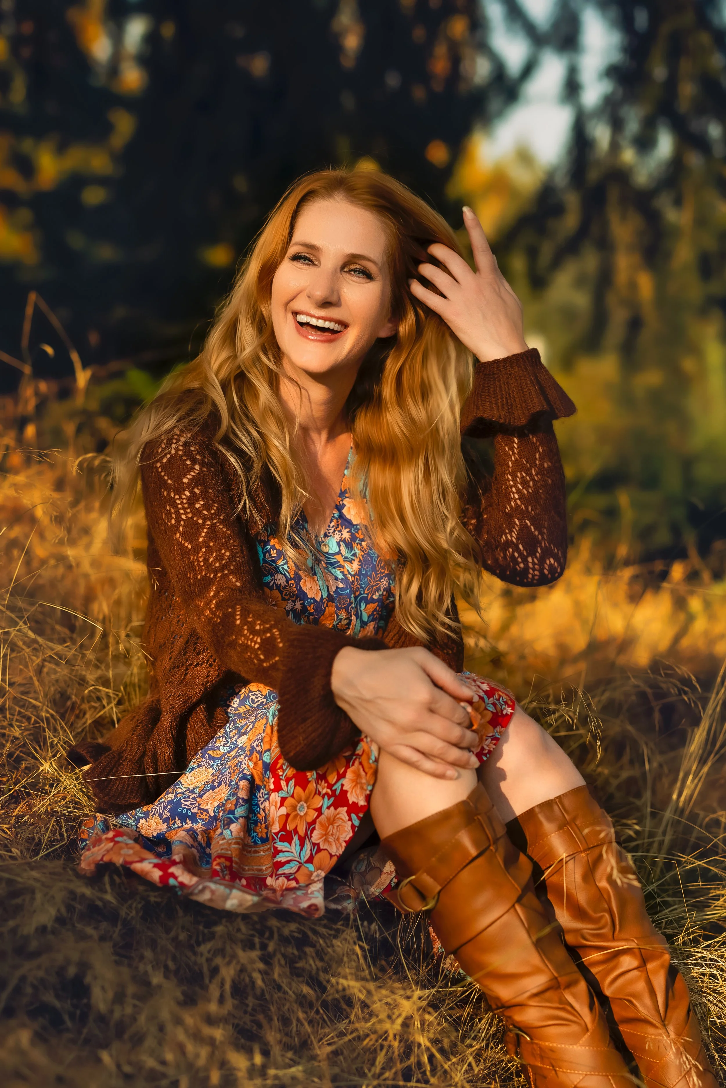 A woman with long wavy blonde hair sitting outdoors in a field during sunset, smiling, wearing a floral dress, a brown knit cardigan, and knee-high brown boots.