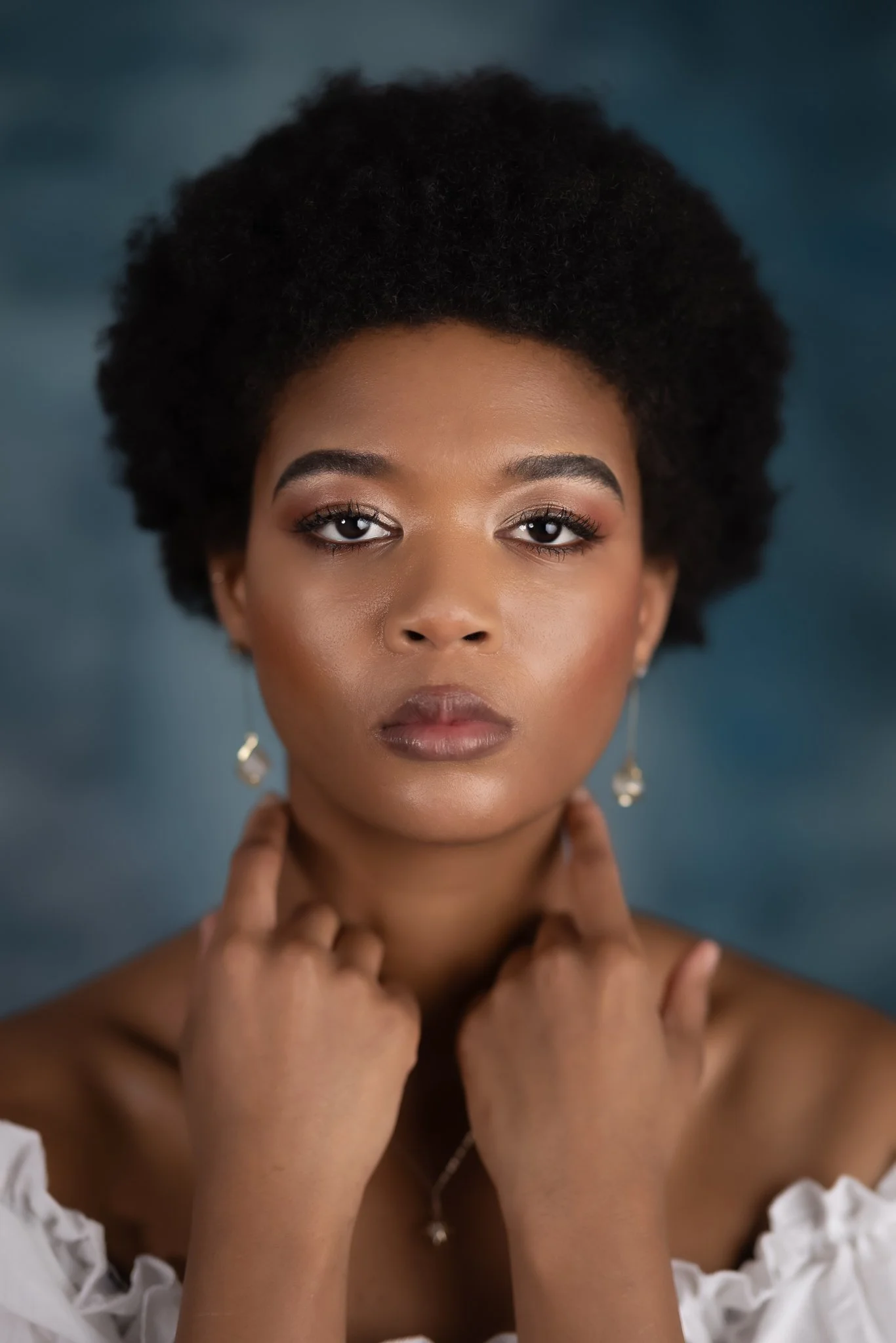 Close-up portrait of a young woman with natural curly hair, wearing earrings and a white off-the-shoulder top, posing against a dark, cloudy background.