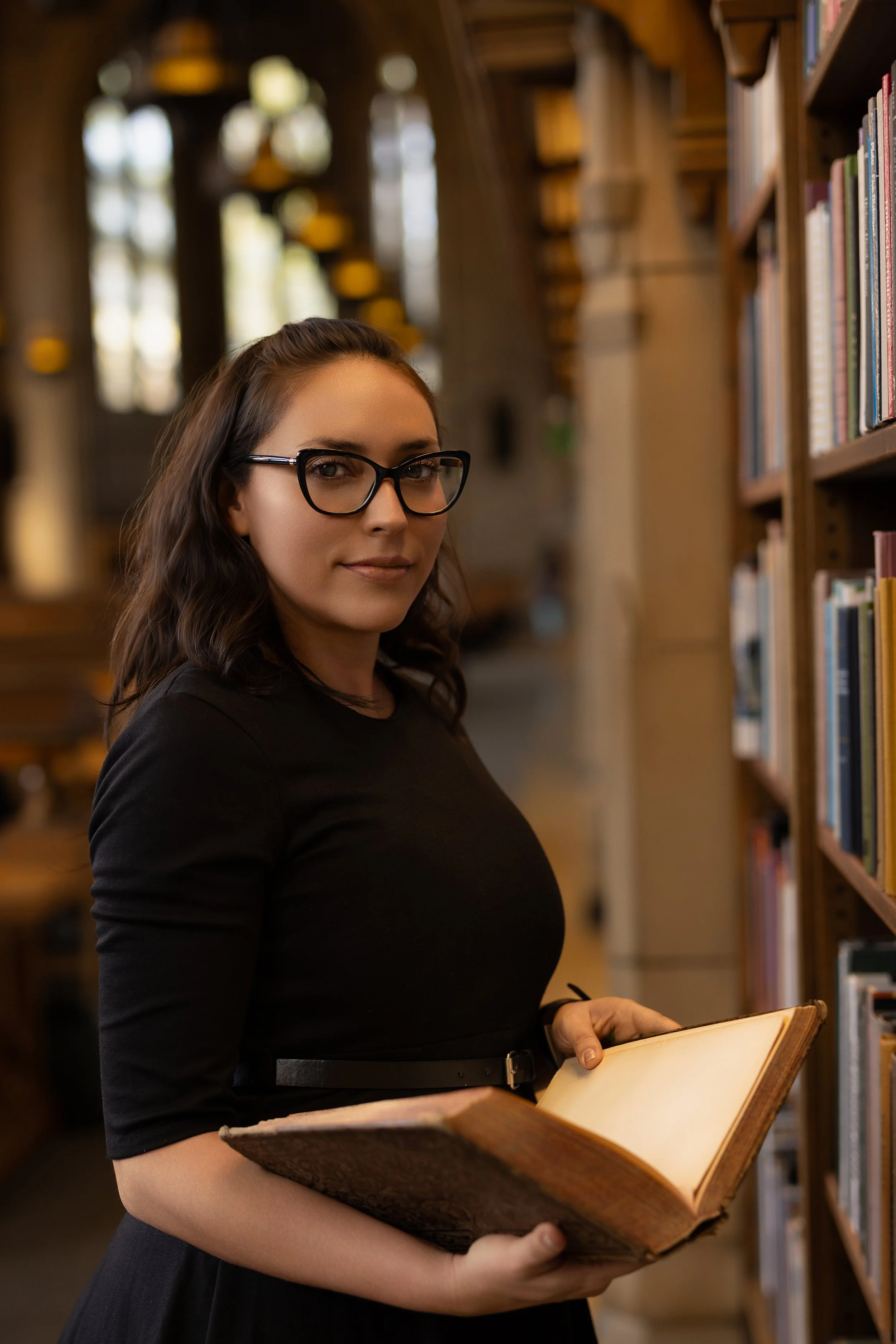 A woman with dark hair, glasses, and a black dress holding an open book in a library.