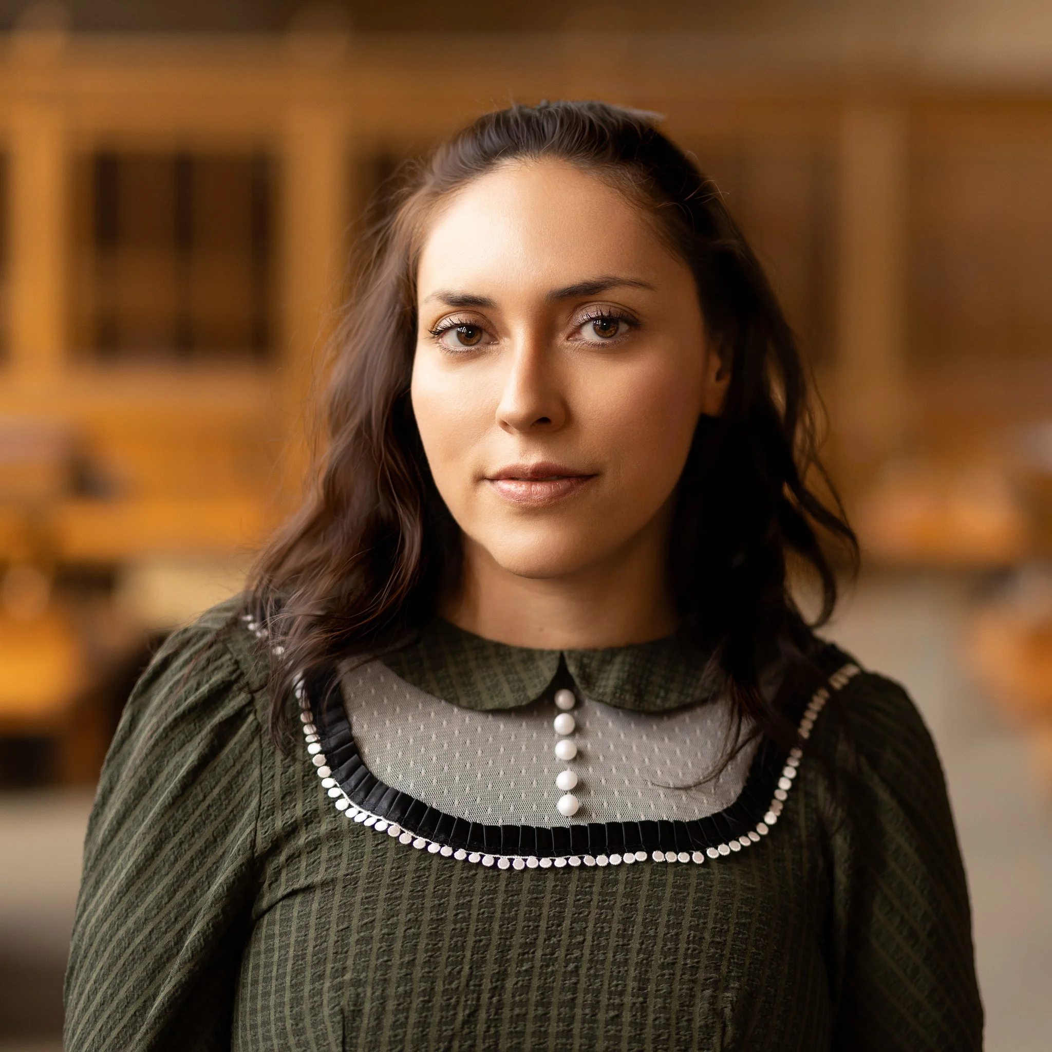 A woman with long dark hair, light skin, and wearing a green dress with a decorative collar, standing in a room with wooden shelves in the background.