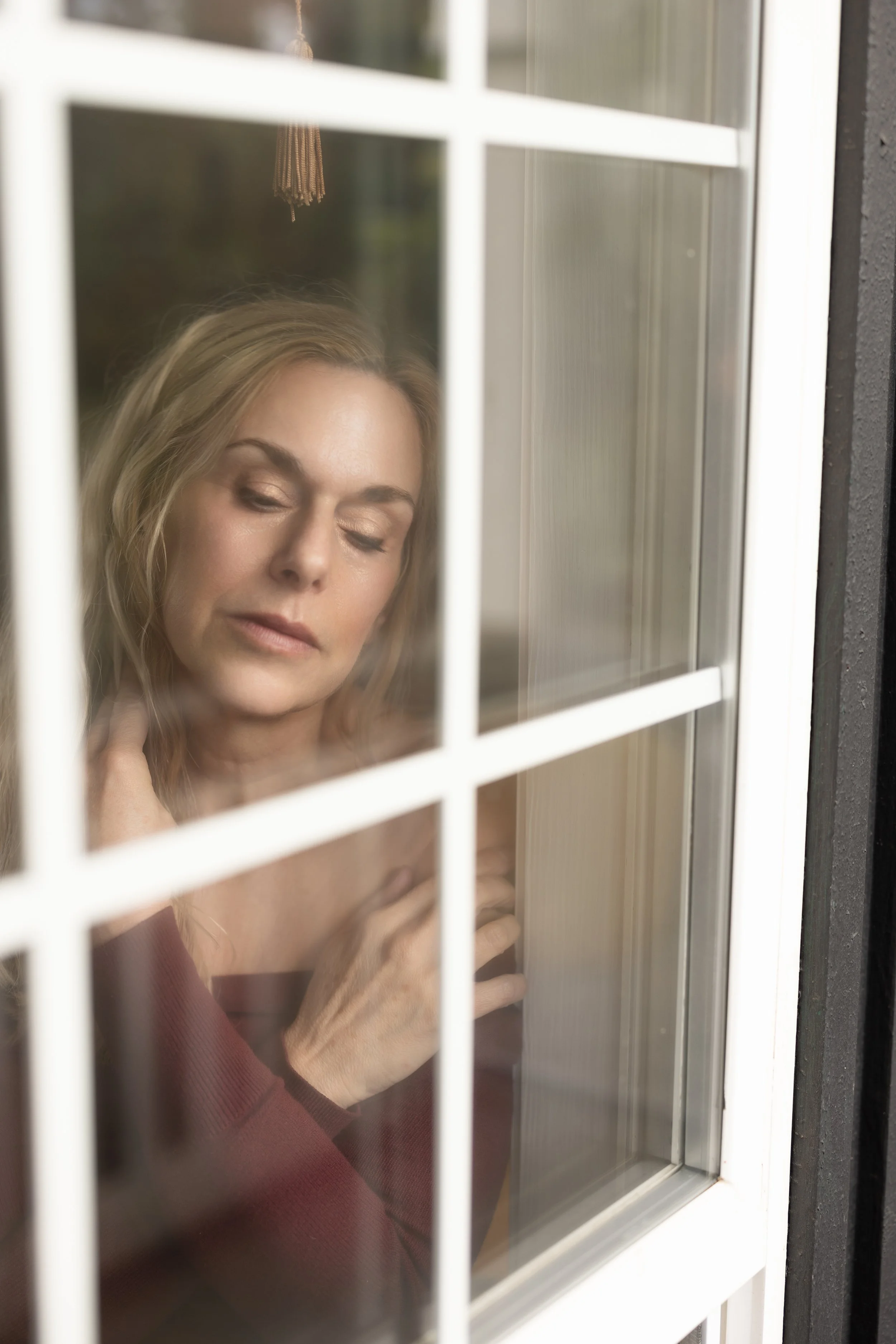 A woman seen through a window with divided panes, touching her neck, with her eyes closed and a calm expression.