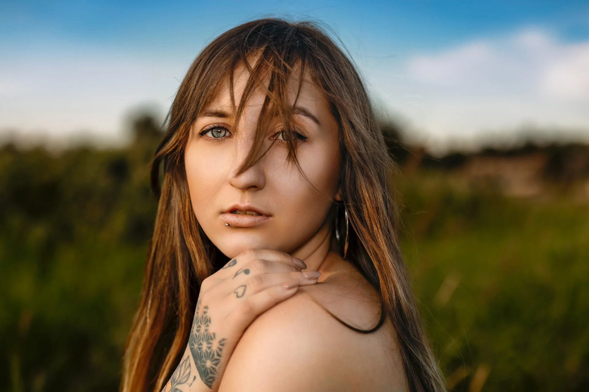 A young woman with long brown hair, tattoos on her left hand, and a piercing on her lower lip, looking at the camera with a landscape background.