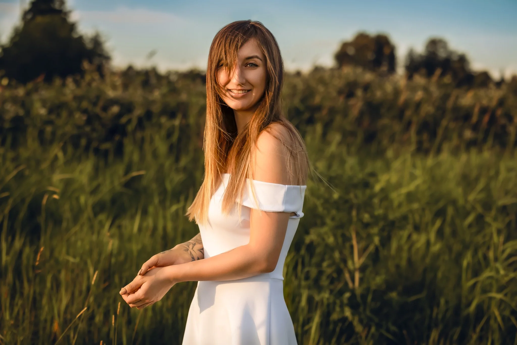 A young woman with long red hair smiling outdoors in a white off-shoulder dress, standing in a green field during sunset.