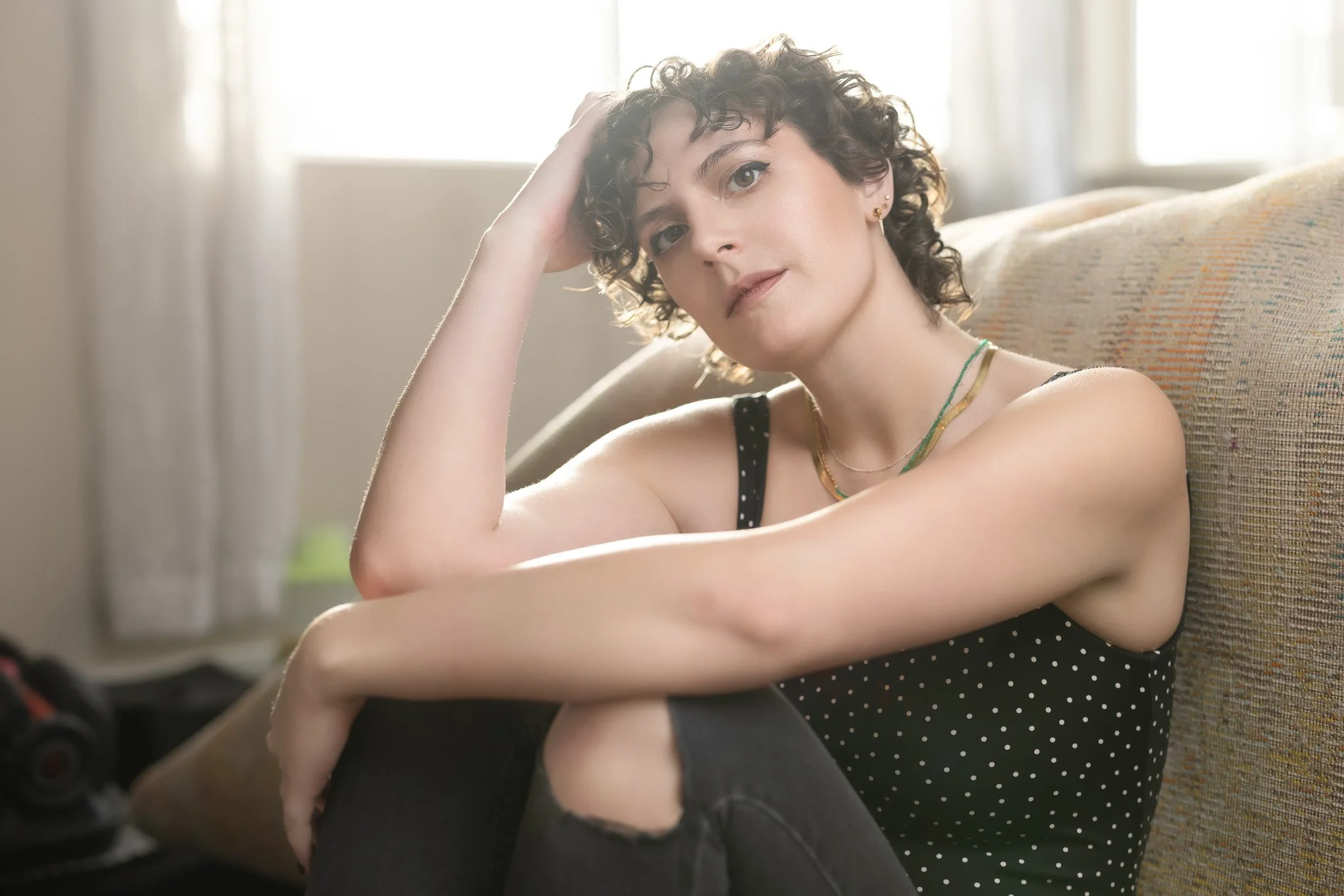 A woman with short curly hair and light skin sitting on a beige couch, wearing a black polka dot tank top and ripped black jeans, looking at the camera with her head resting on her hand.