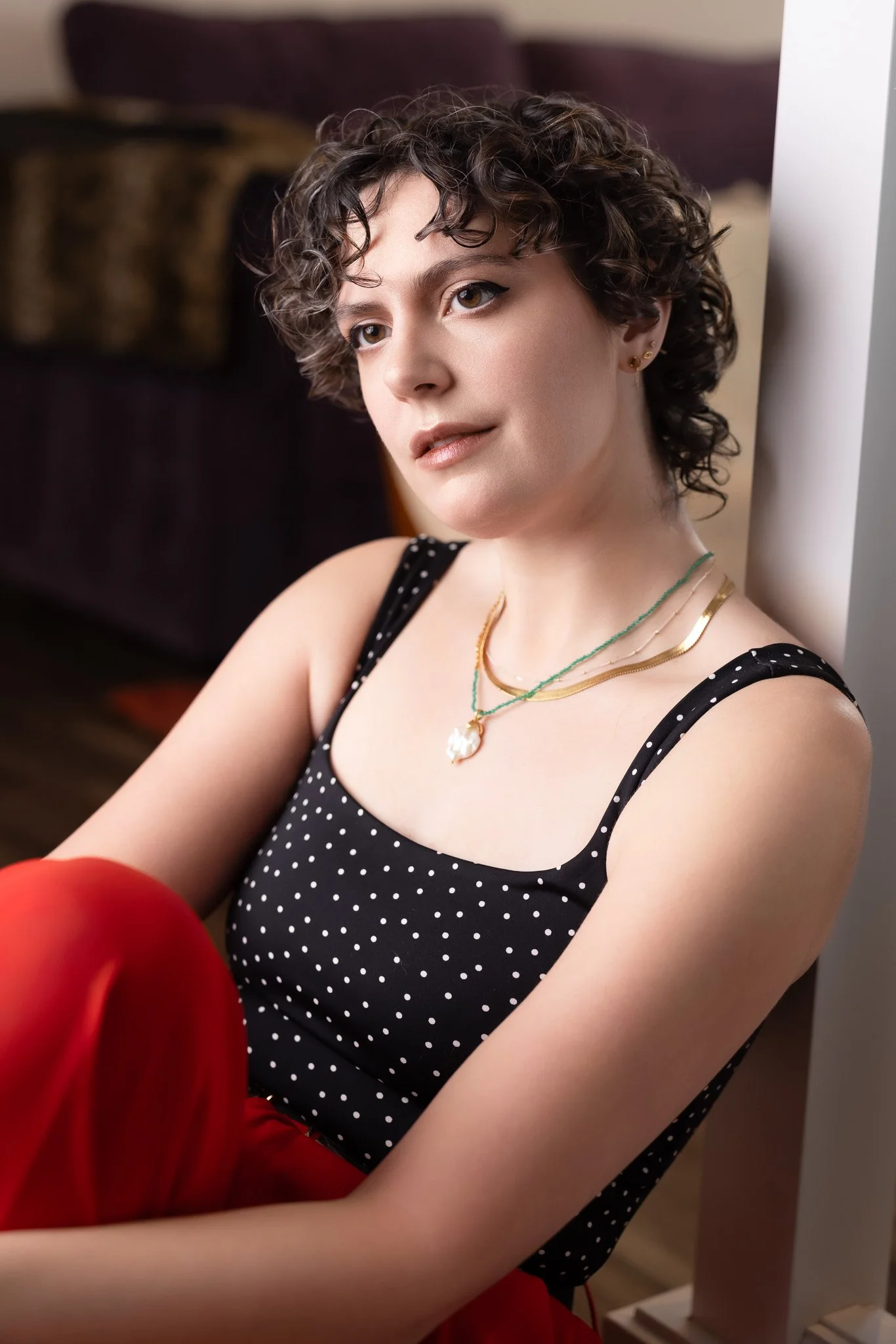 A young woman with short, curly hair sitting against a wall, wearing a black polka dot top and red pants, accessorized with layered necklaces and small earrings.