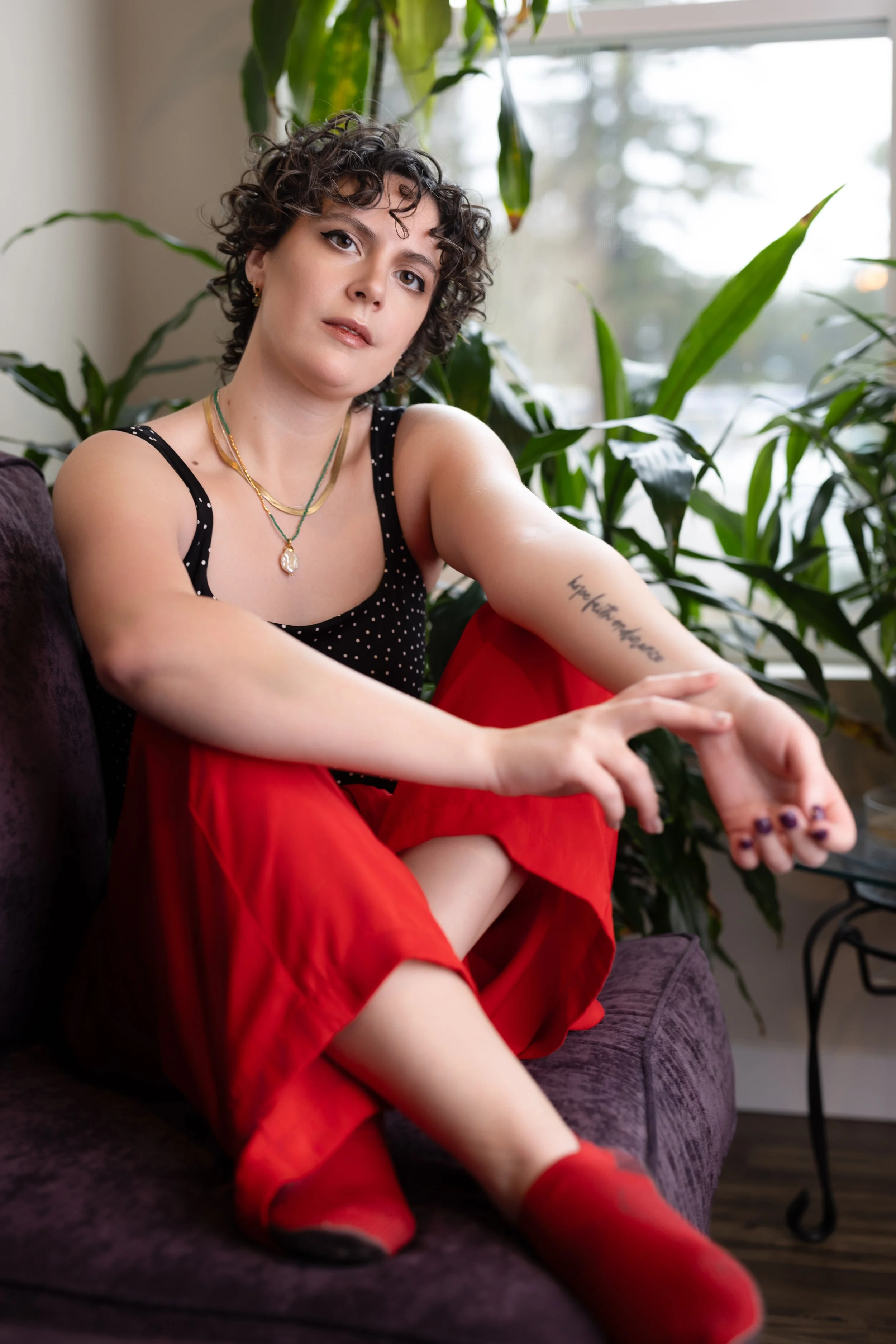 A woman with short curly hair, wearing a black polka dot top, red skirt, and red socks, sits on a purple couch near a window surrounded by green plants.