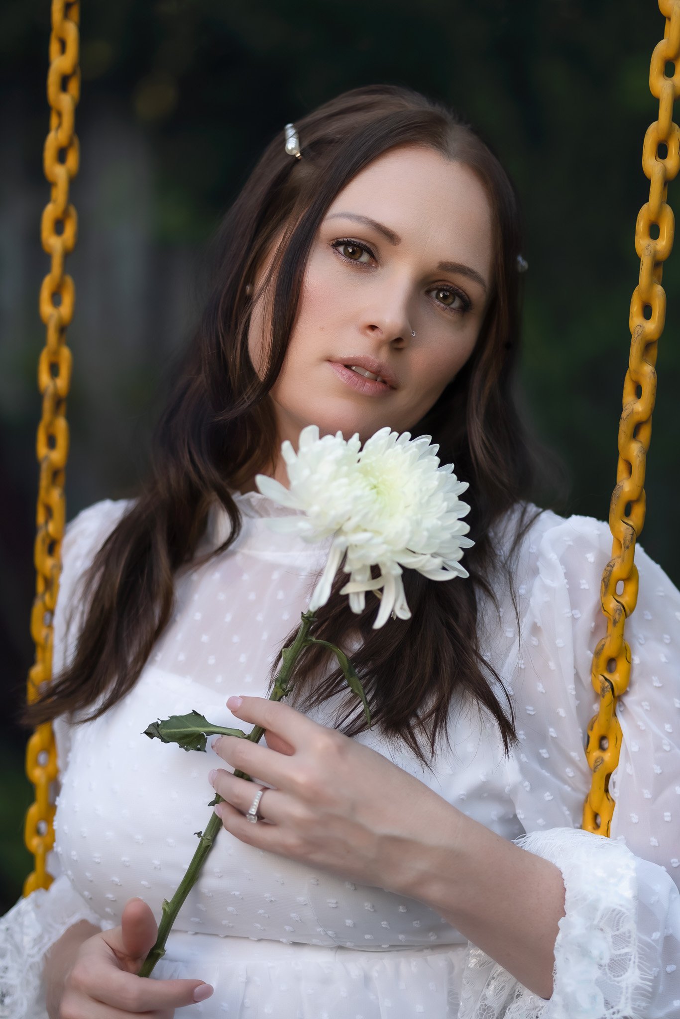 A woman with dark brown hair and a white blouse holding a white chrysanthemum flower, sitting on a yellow swing outdoors.