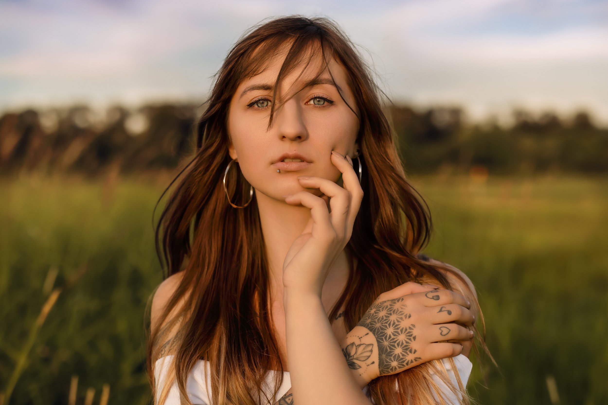 A young woman with long reddish-brown hair, piercing blue eyes, and multiple tattoos, standing in a grassy field during sunset, looking directly at the camera with her hand near her chin.