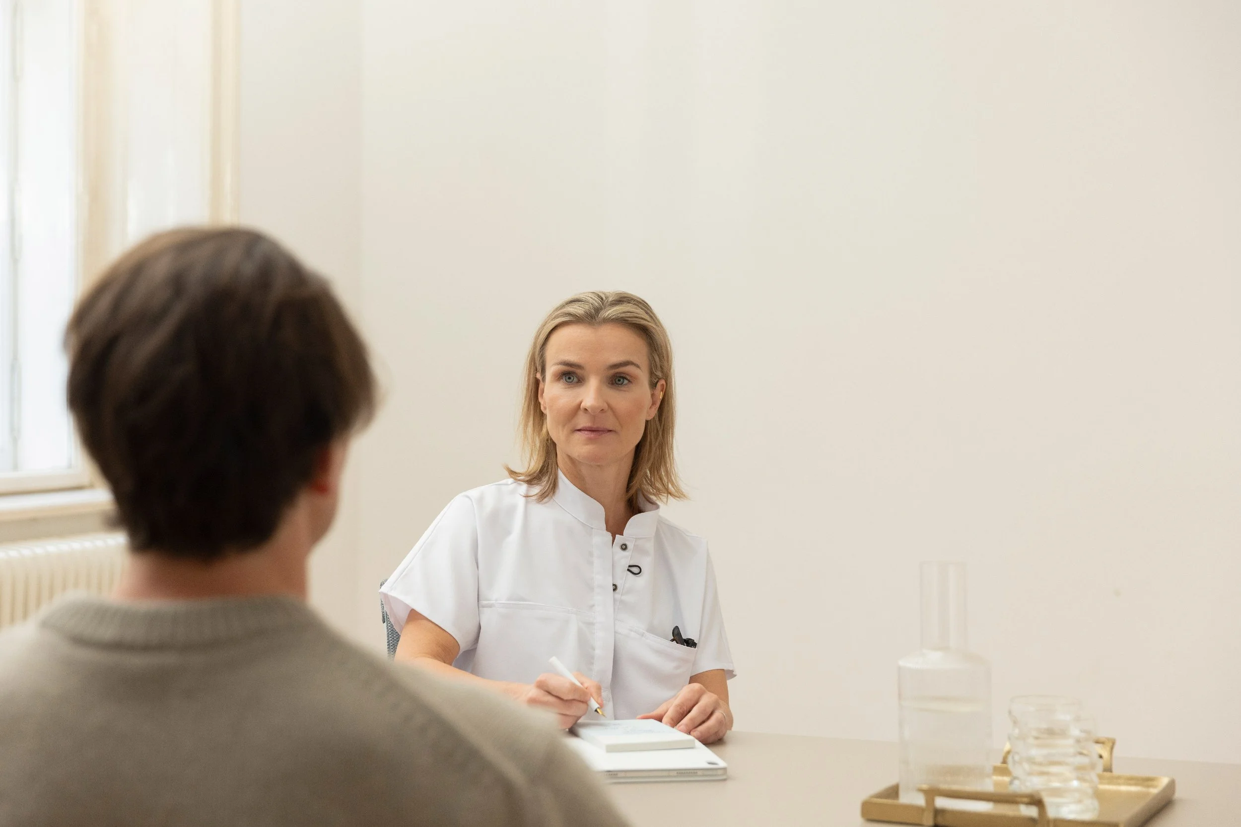 Ärztin im Gespräch mit Patient in einer Praxis, schreibt Notizen. Hintergrund ist eine weiße Wand, es stehen Wasserflaschen und Gläser auf einem Tablett.