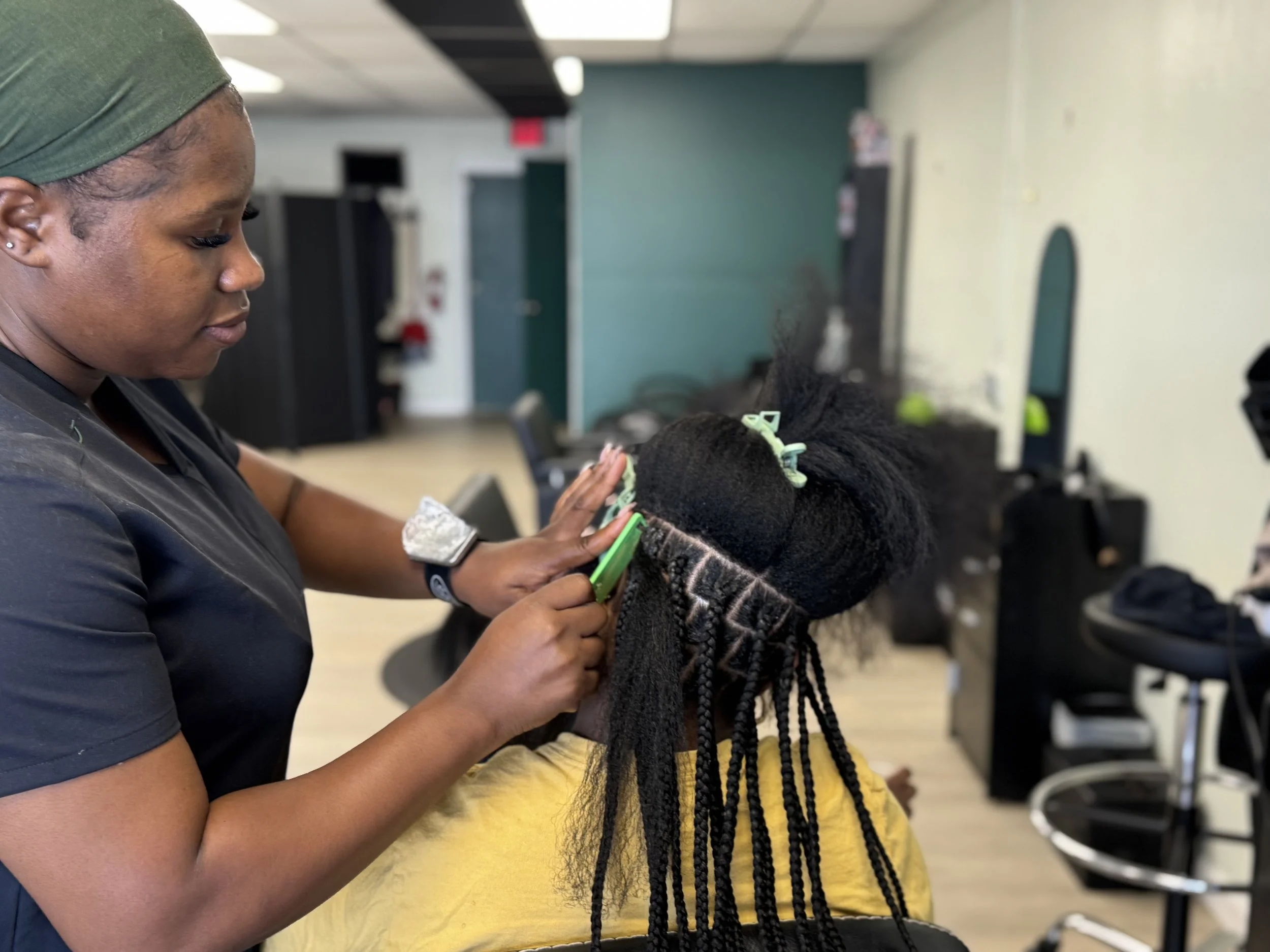 A stylist trimming a woman's braided hair in a salon, with hair clips, scissors, and salon furniture visible.
