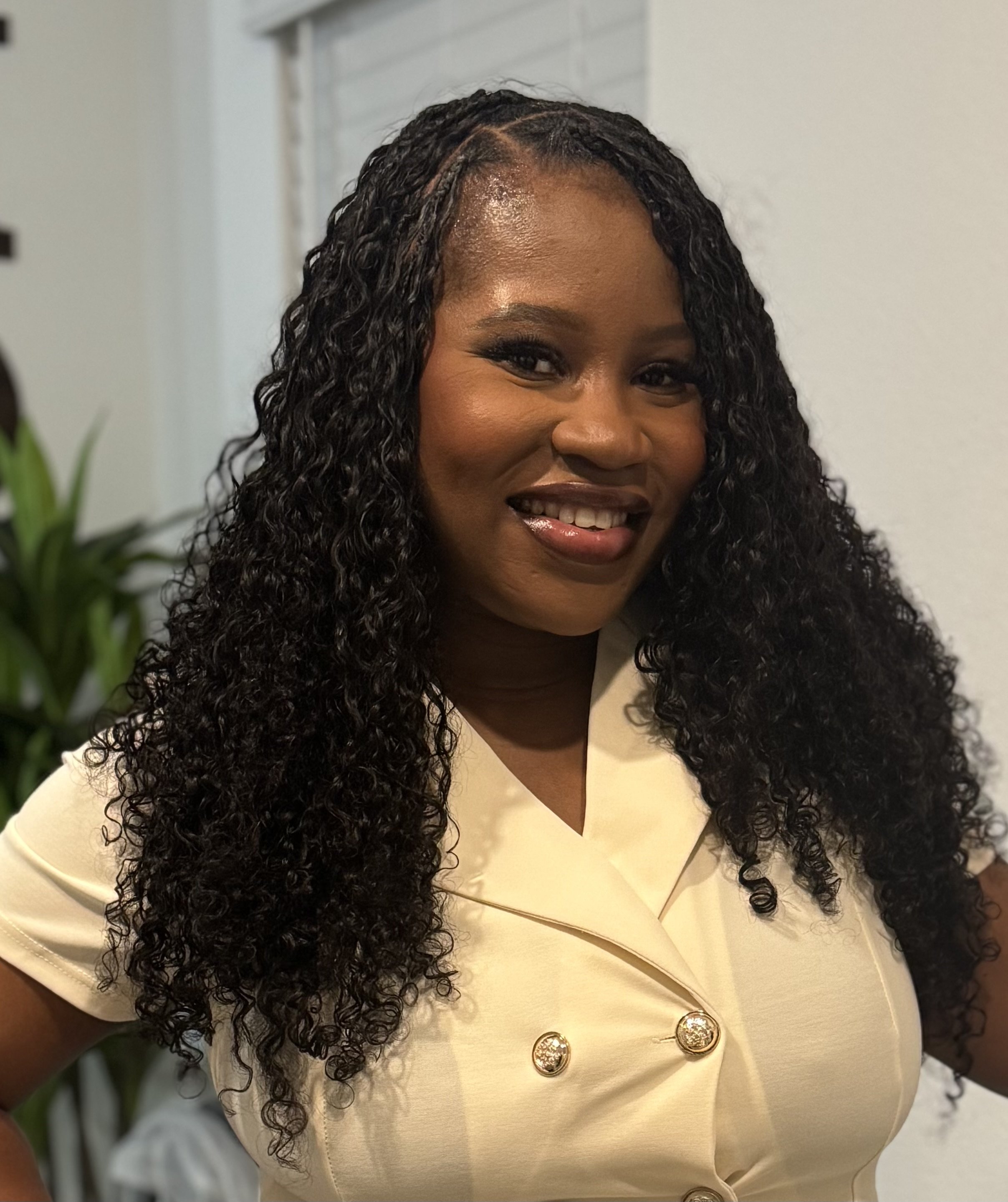 A woman with long curly black hair smiling, wearing a cream-colored outfit with gold buttons, standing indoors near a white wall and a green plant.