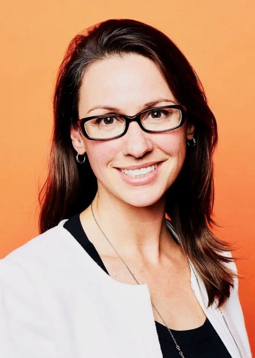 A woman with long brown hair, glasses, and earrings, smiling against an orange background.