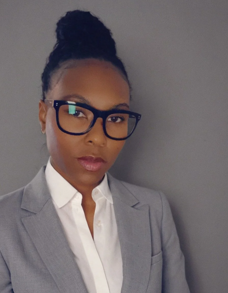 A woman wearing glasses and a light gray blazer over a white shirt, standing against a plain gray wall.