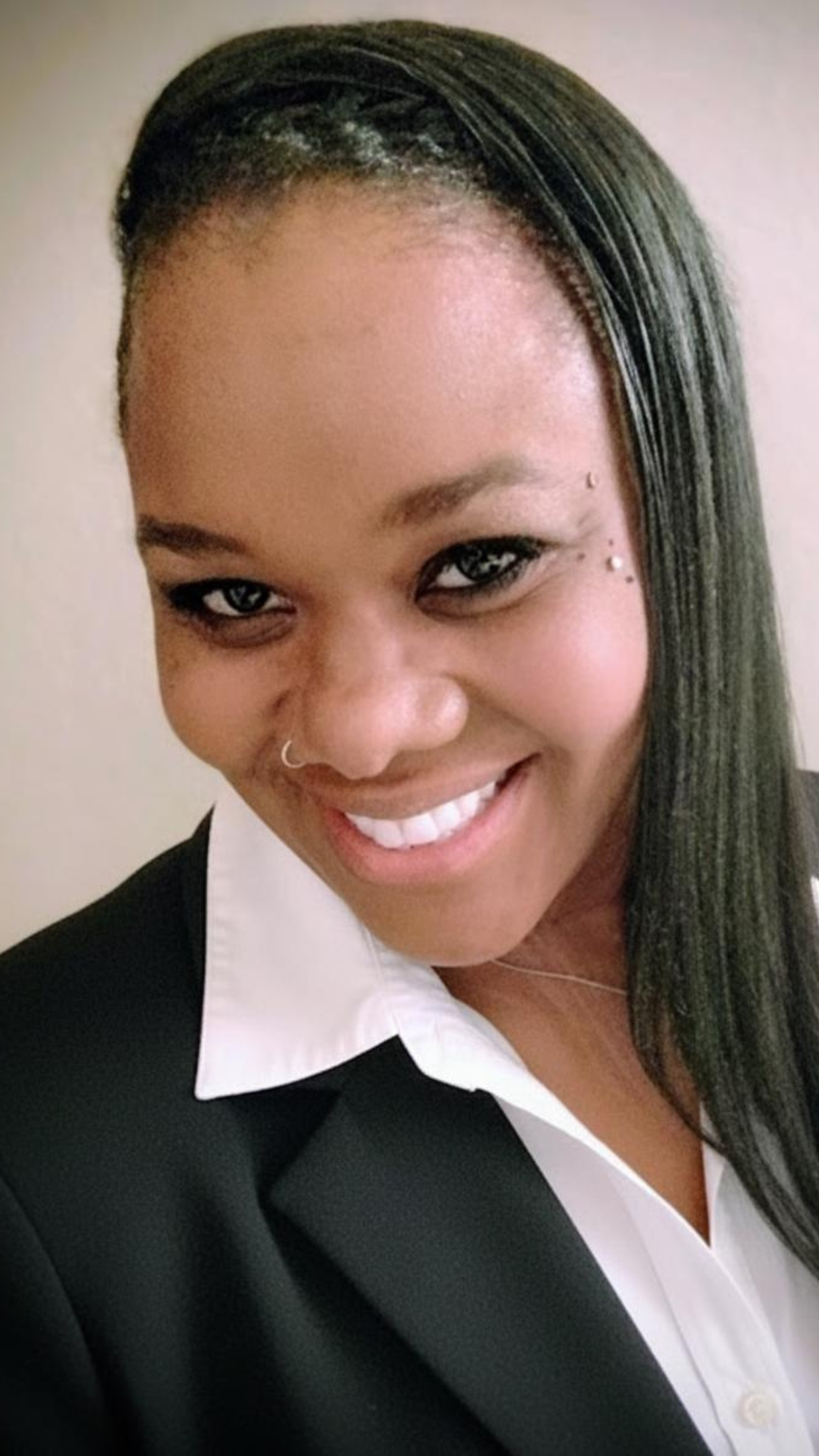 A woman smiling with a side glance, wearing a black blazer over a white shirt, with earrings and a nose ring.